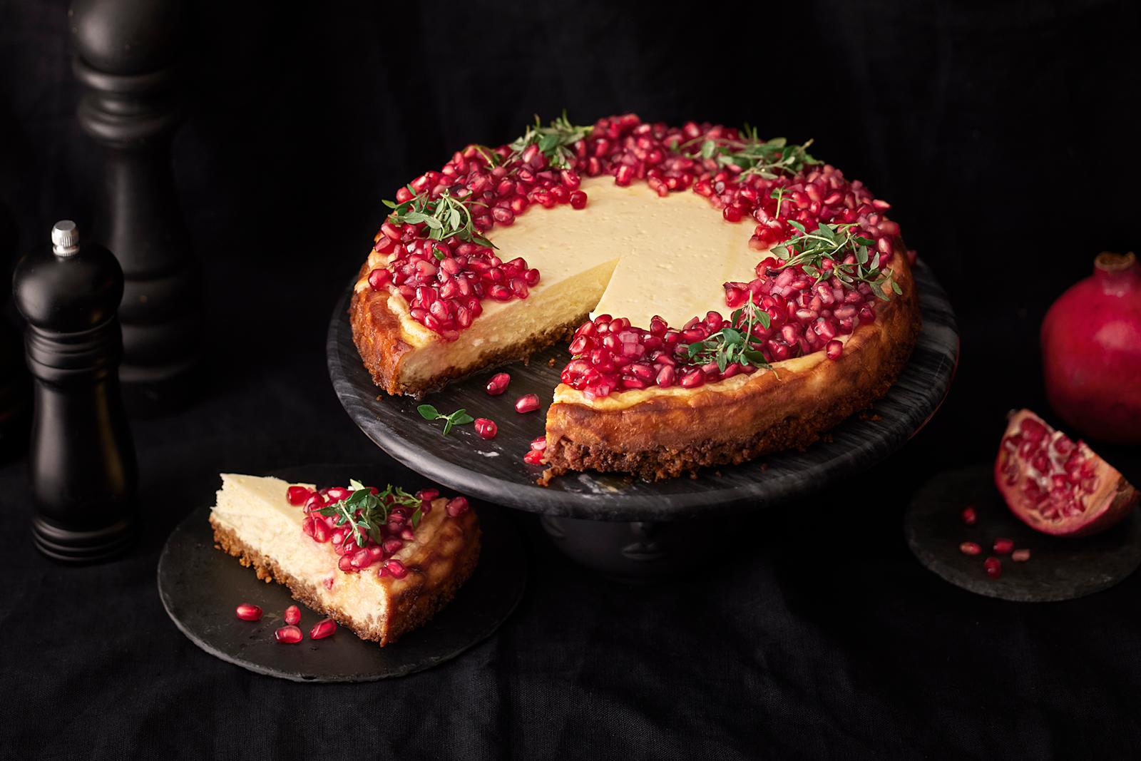 Granaattiomenasalsalla koristeltu suolainen juustokakku, josta leikattu pala lautaselle. Kakun vierellä granattiomenalohko ja kokonainen granaattiomena sekä kolme tummaa pippurimyllyä.