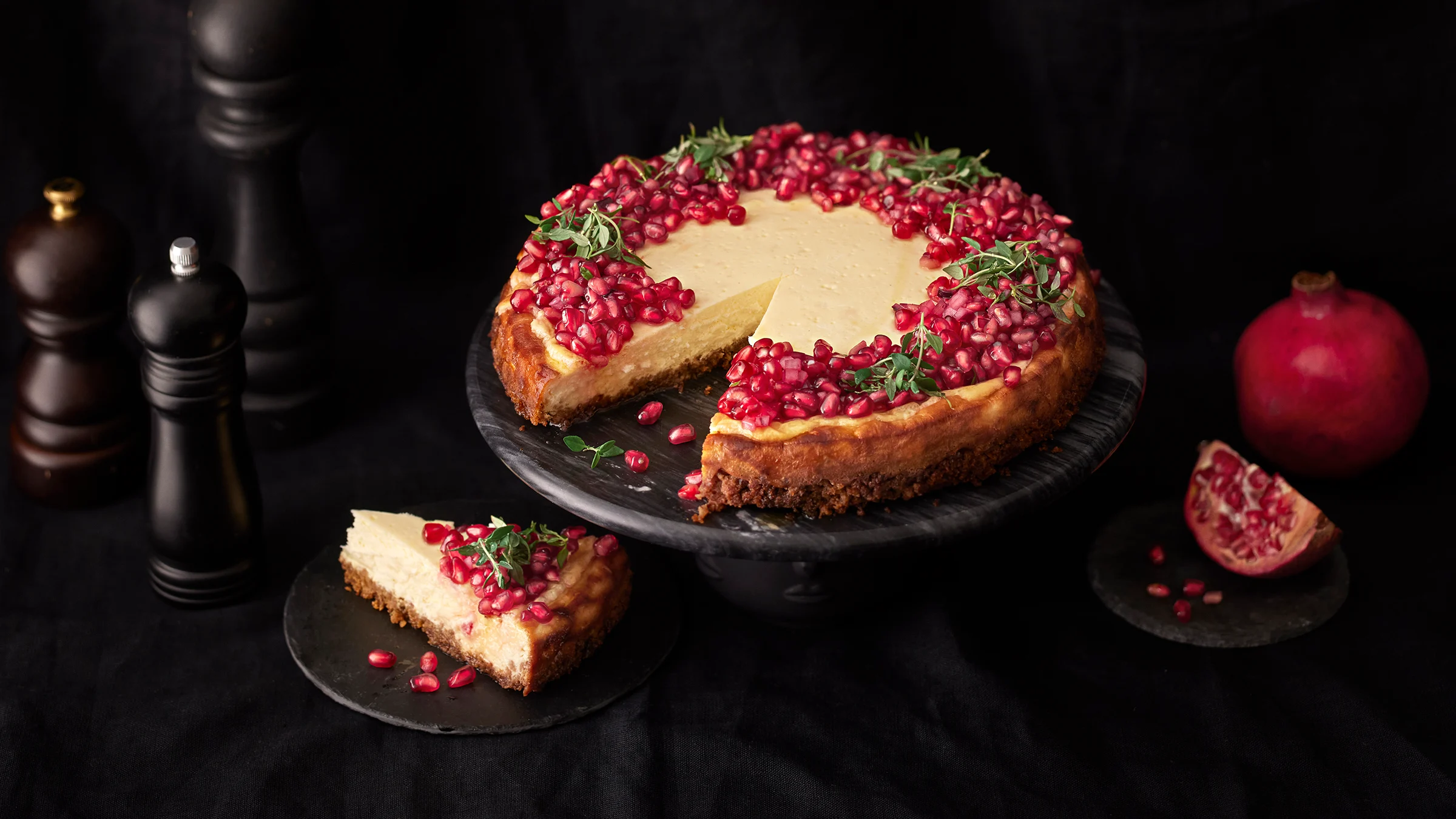 Granaattiomenasalsalla koristeltu suolainen juustokakku, josta leikattu pala lautaselle. Kakun vierellä granattiomenalohko ja kokonainen granaattiomena sekä kolme tummaa pippurimyllyä.