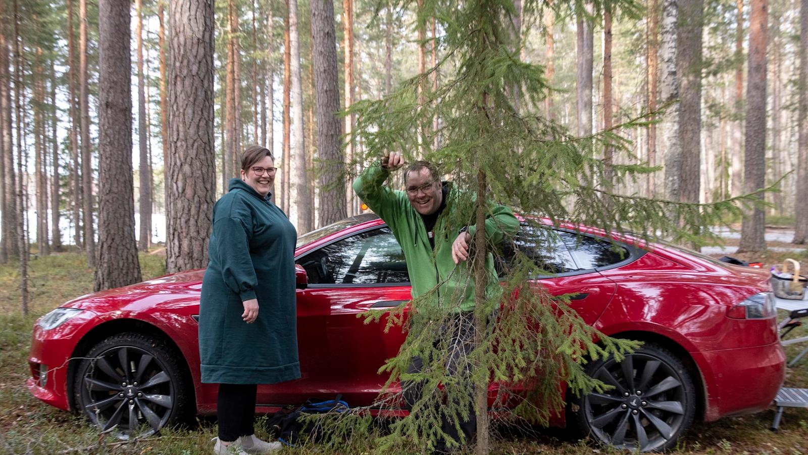 Katja ja Sami Tuomola seisovat Teslan edessä suomalaisessa metsässä.