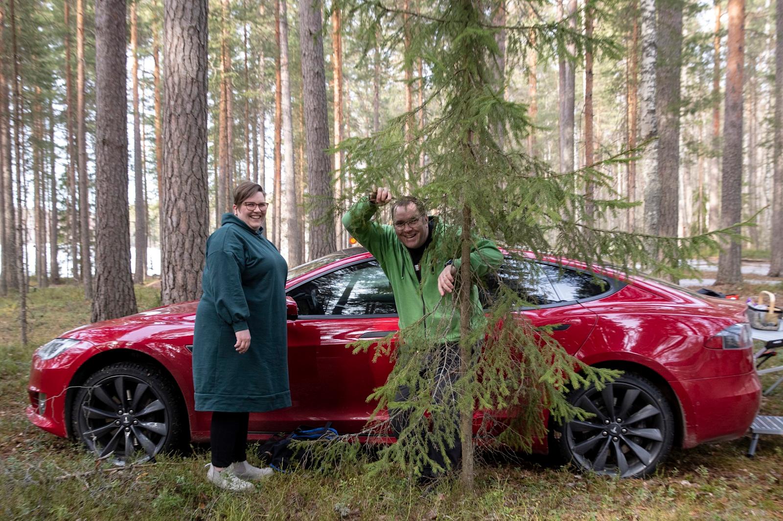 Katja ja Sami Tuomola seisovat Teslan edessä suomalaisessa metsässä.