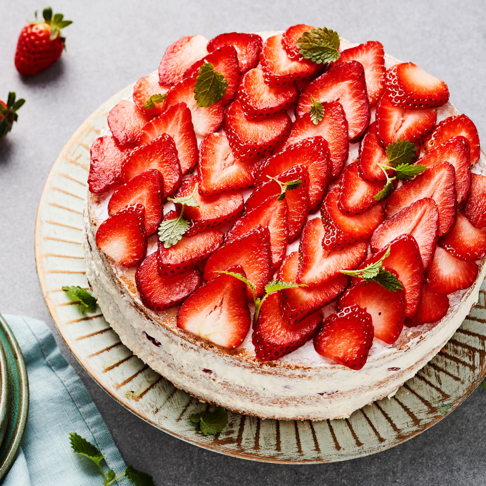 Mansikkakakku, joka on koristeltu viipaloiduilla mansikoilla. Vieressä mansikoita, mansikan lehtiä ja pino lautasia.