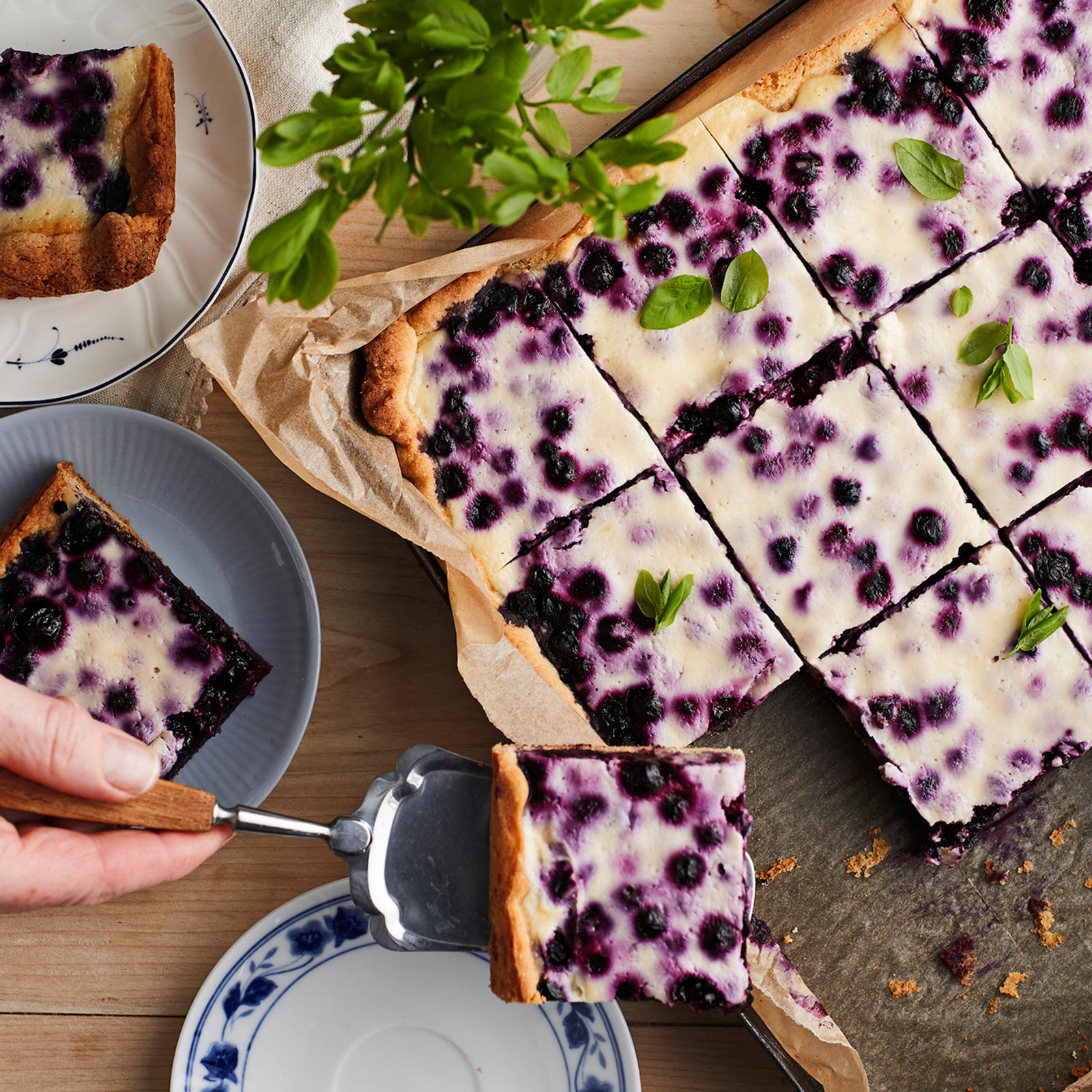 Pellillinen paloiksi leikattua mustikkapiirakkaa, kaksi lautasta joilla piirakkapala sekä yksi tyhjä lautanen, jolle ollaan nostamassa piirakkapalaa kakkulapion avulla.