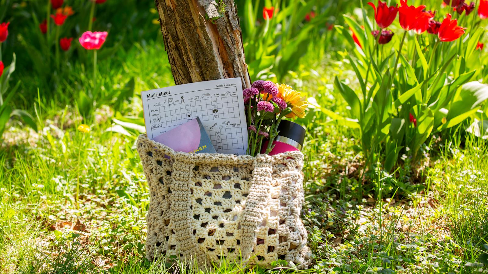 Virkattu kesäkassi, jonka sisällä ristikkolehti, kirja, kukkia ja juomapullo.