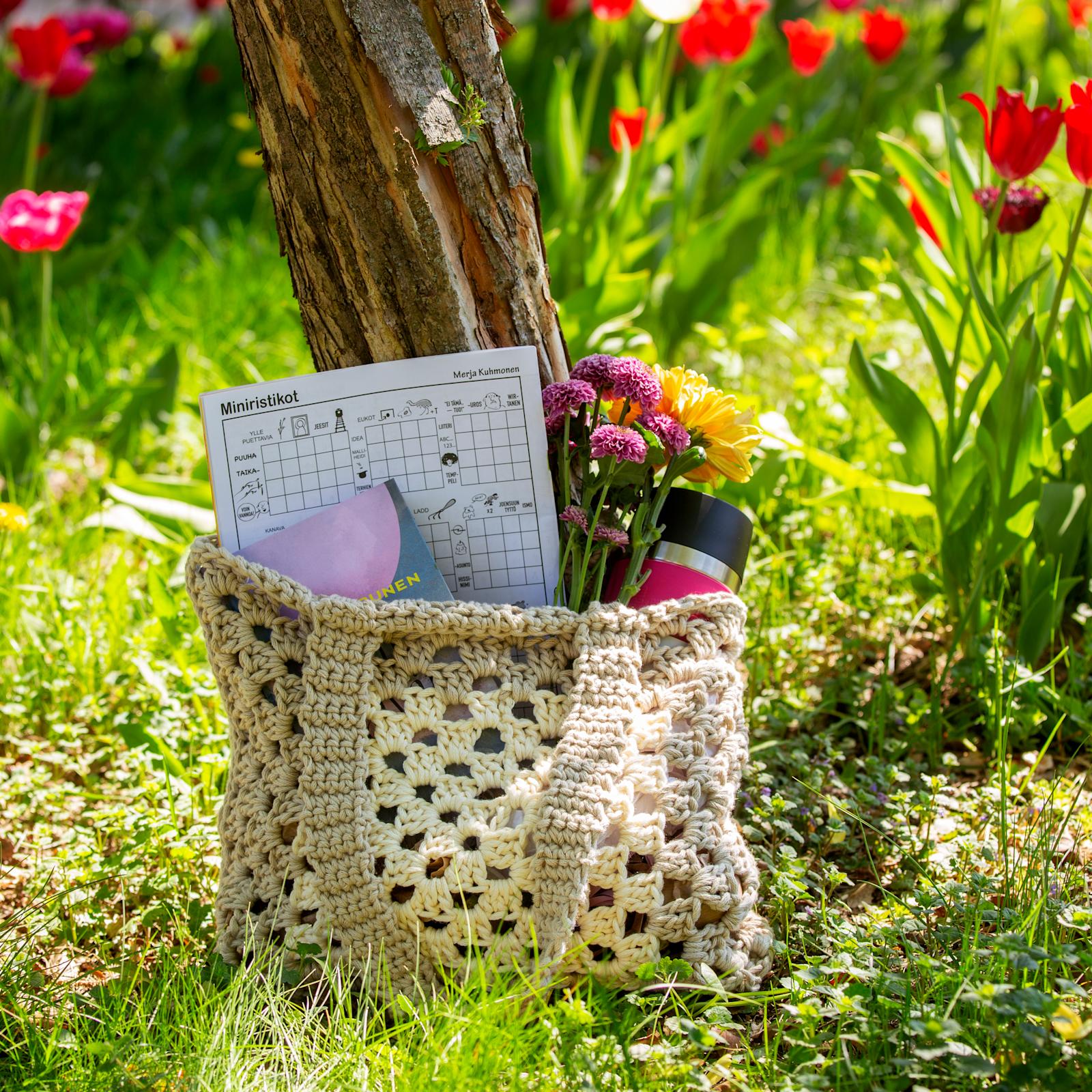 Virkattu kesäkassi, jonka sisällä ristikkolehti, kirja, kukkia ja juomapullo.