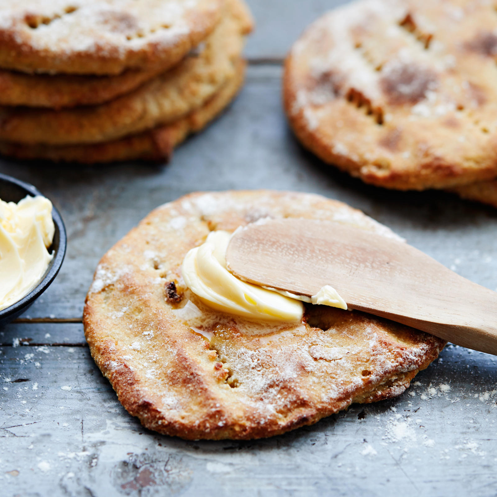 Perunarieska, jonka päällä voita ja puinen voiveitsi. Vieressä pieni kulhollinen voita sekä kaksi pinoa perunarieskoja.