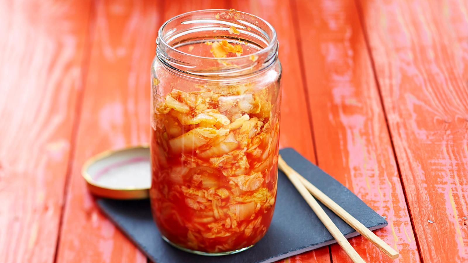 Pikakimchiä korkeassa lasipurkissa. Purkin alla musta nelikulmainen lautanen, jonka päällä syömäpuikot ja purkin kansi.