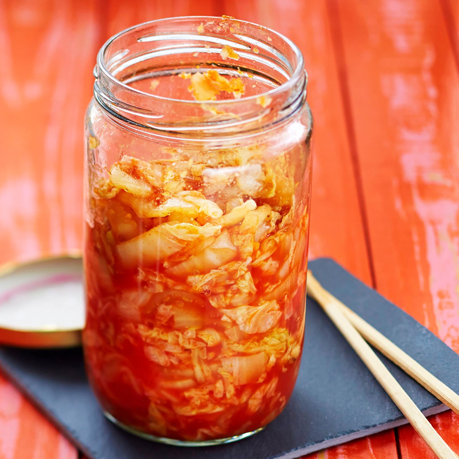 Pikakimchiä korkeassa lasipurkissa. Purkin alla musta nelikulmainen lautanen, jonka päällä syömäpuikot ja purkin kansi.