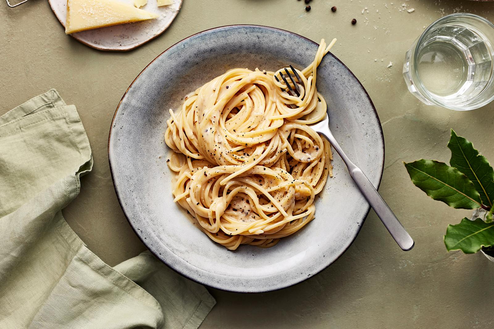 Cacio e pepe -pastaa lautasella. Annoksen vieressä vesilasi, vihreä liina, pecorinojuustoa, pippureita ja kasvin lehtiä.