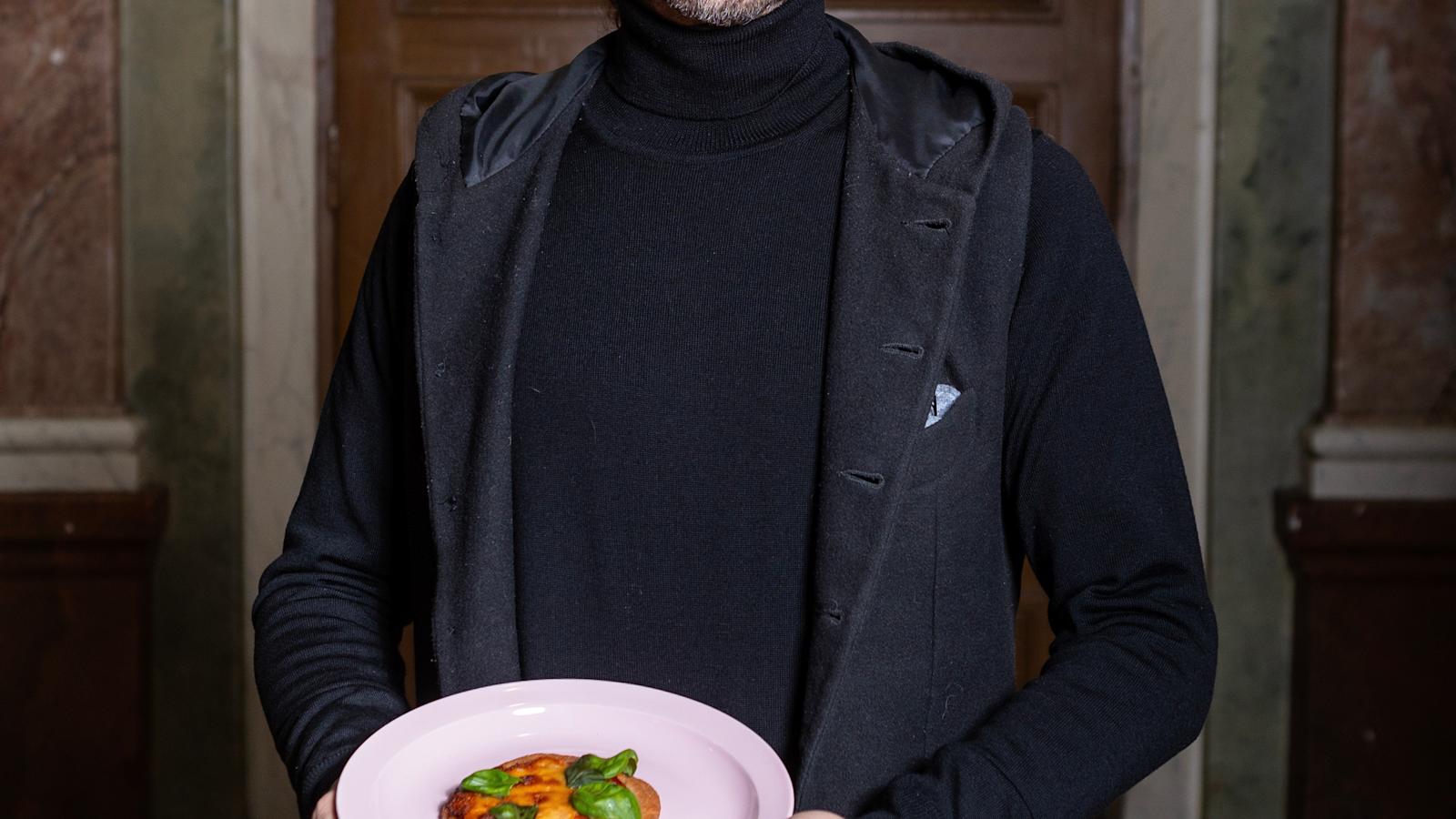 Jarmo Lipasti on pukeutunut mustaan pooloneuleeseen ja harmaaseen liiviin. Hän pitelee kädessään valkoista lautasta, jolla on pieni pizzapalanen.