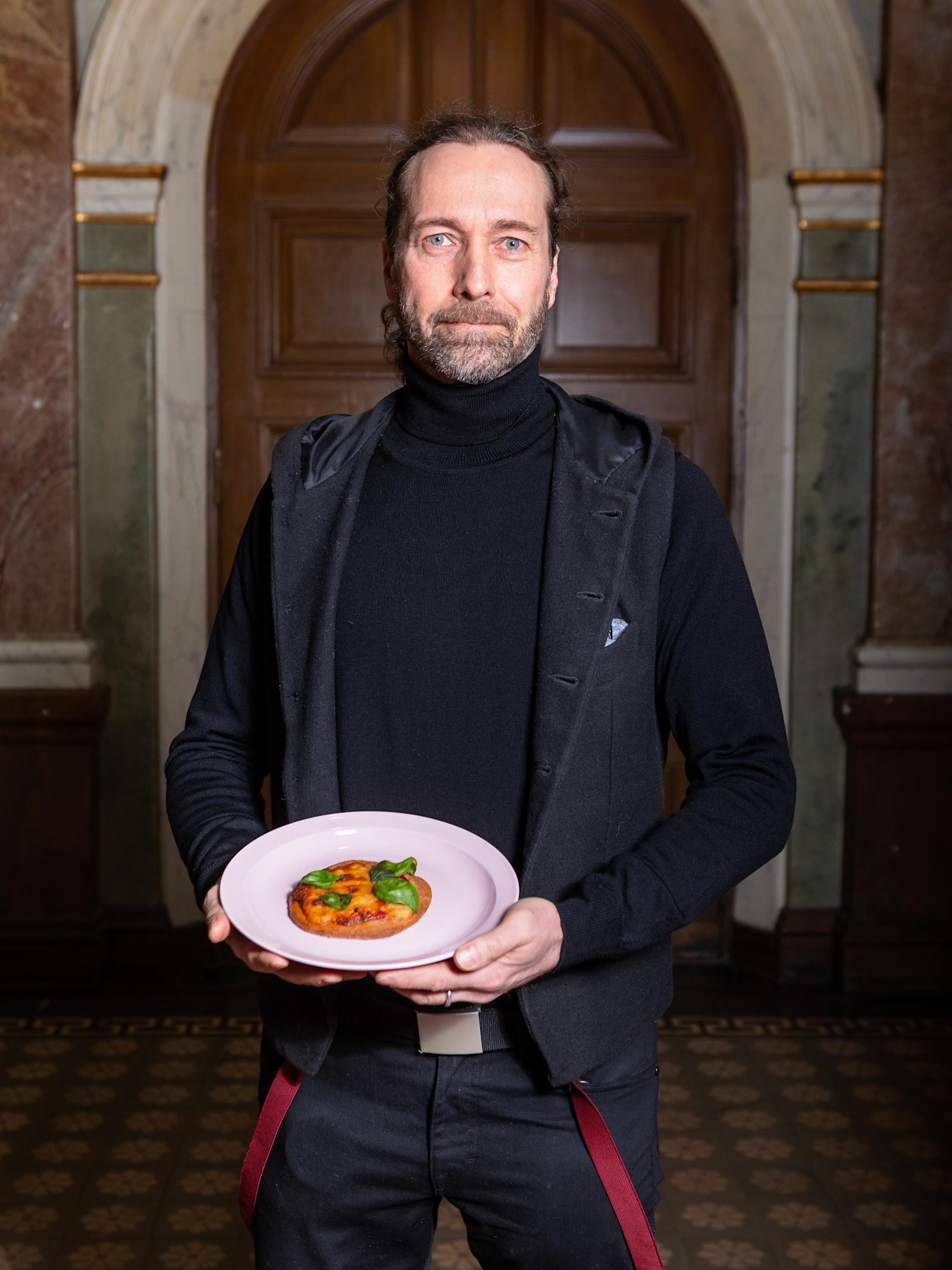 Jarmo Lipasti on pukeutunut mustaan pooloneuleeseen ja harmaaseen liiviin. Hän pitelee kädessään valkoista lautasta, jolla on pieni pizzapalanen.