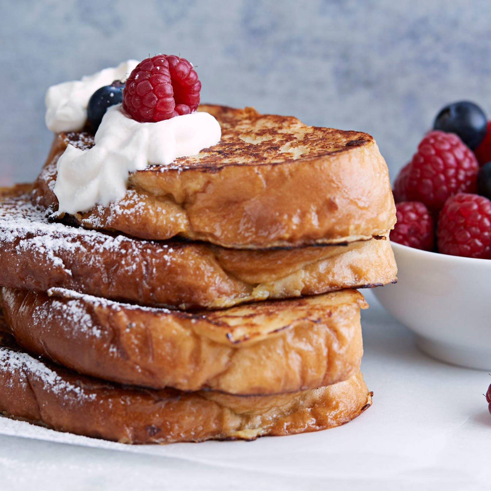 Pino köyhiä ritareita, joiden päällä kermavaahtoa, tuoreita marjoja ja tomusokeria. Sivussa marjoja ja kermavaahtoa kulhoissa.