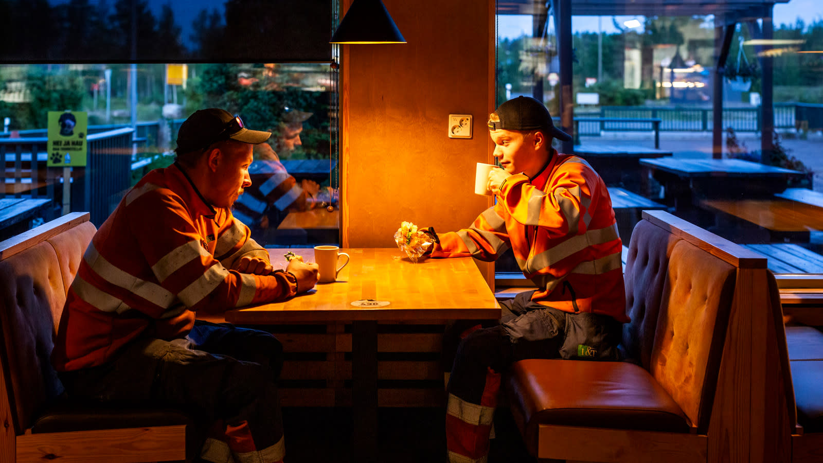 Kaksi oranssiin huomiovaatteisiin ja sinisiin lippiksiin pukeutunutta miestä aamukahvilla huoltoaseman pöydässä.