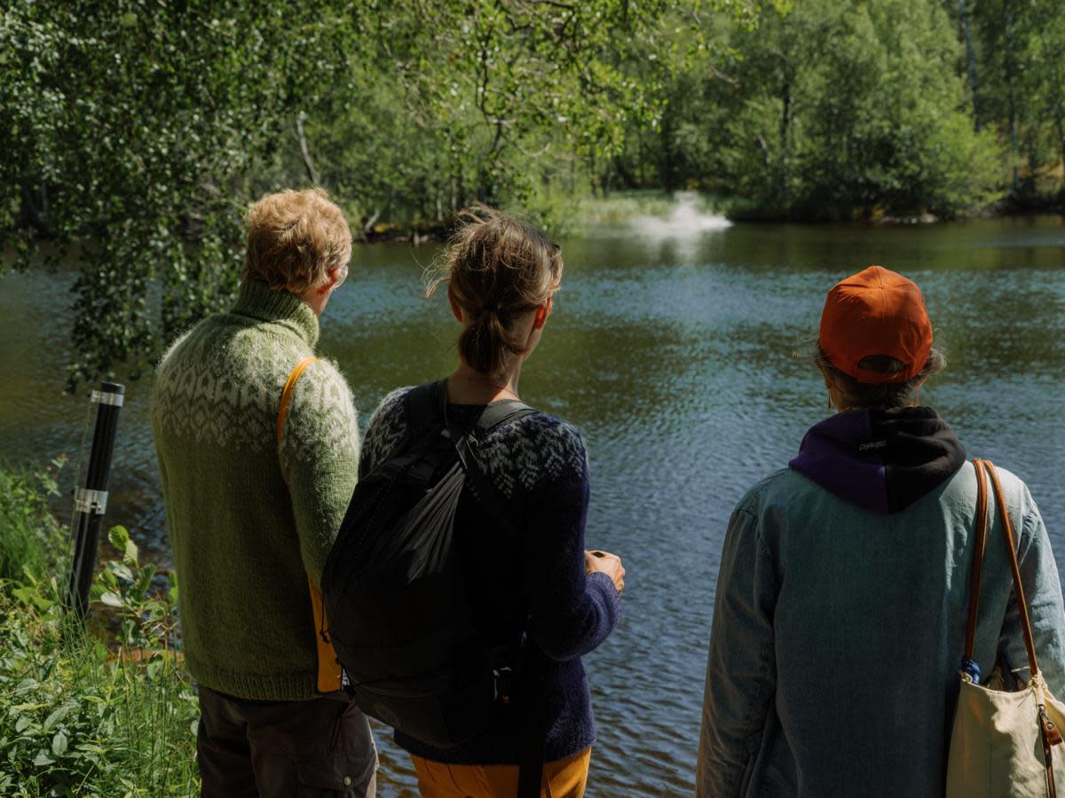 Kolme henkilöä kesäisen lammen rannalla. Lammen pinnalla on utua.