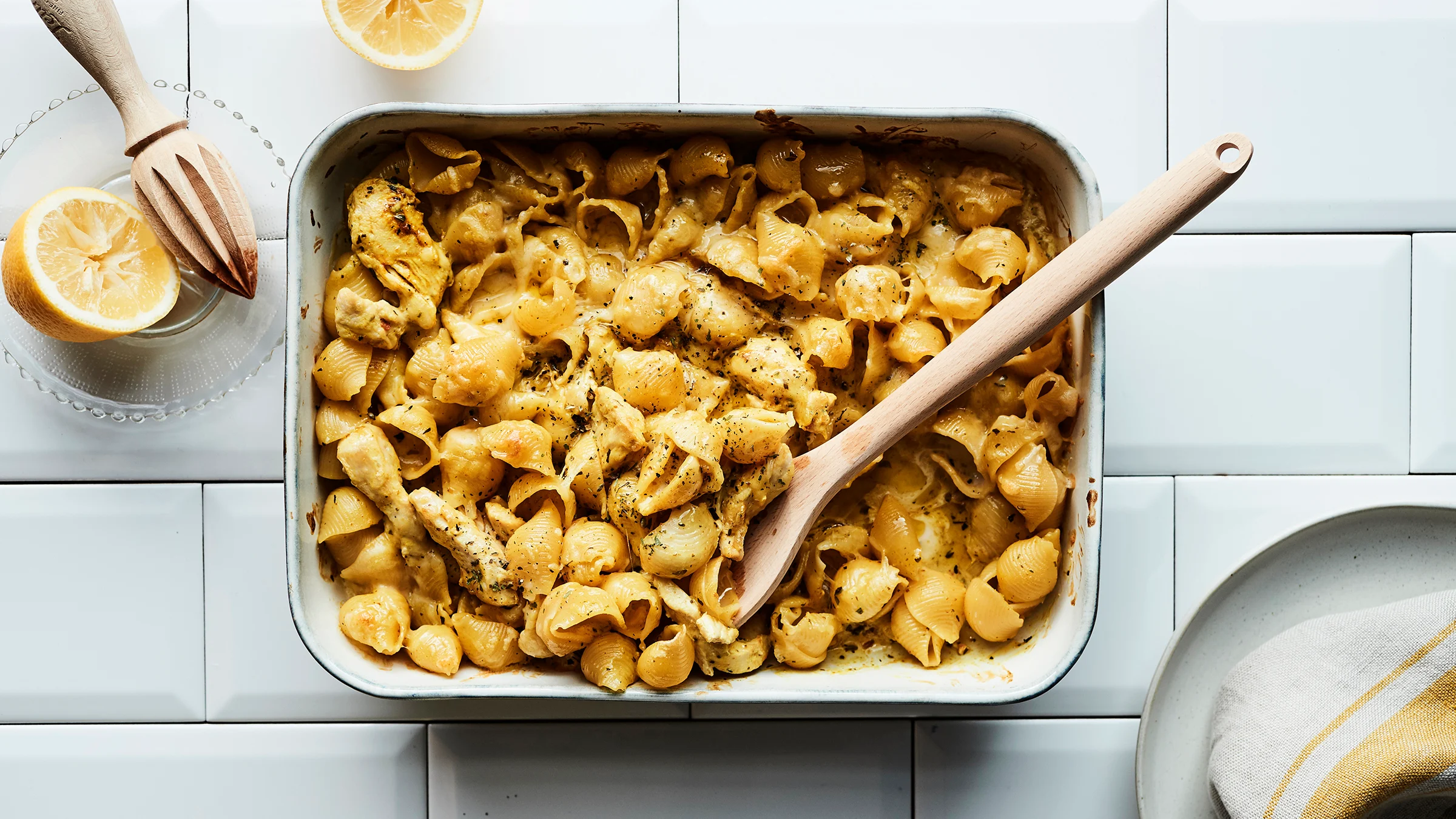 Uunivuoka, jossa Palermon kanapastaa sekä puinen otin. Vieressä puinen sitruspuserrin ja puolikas sitruuna lasilautasella, sekä valkoinen ruokalautanen ja lautasliinan kulma.