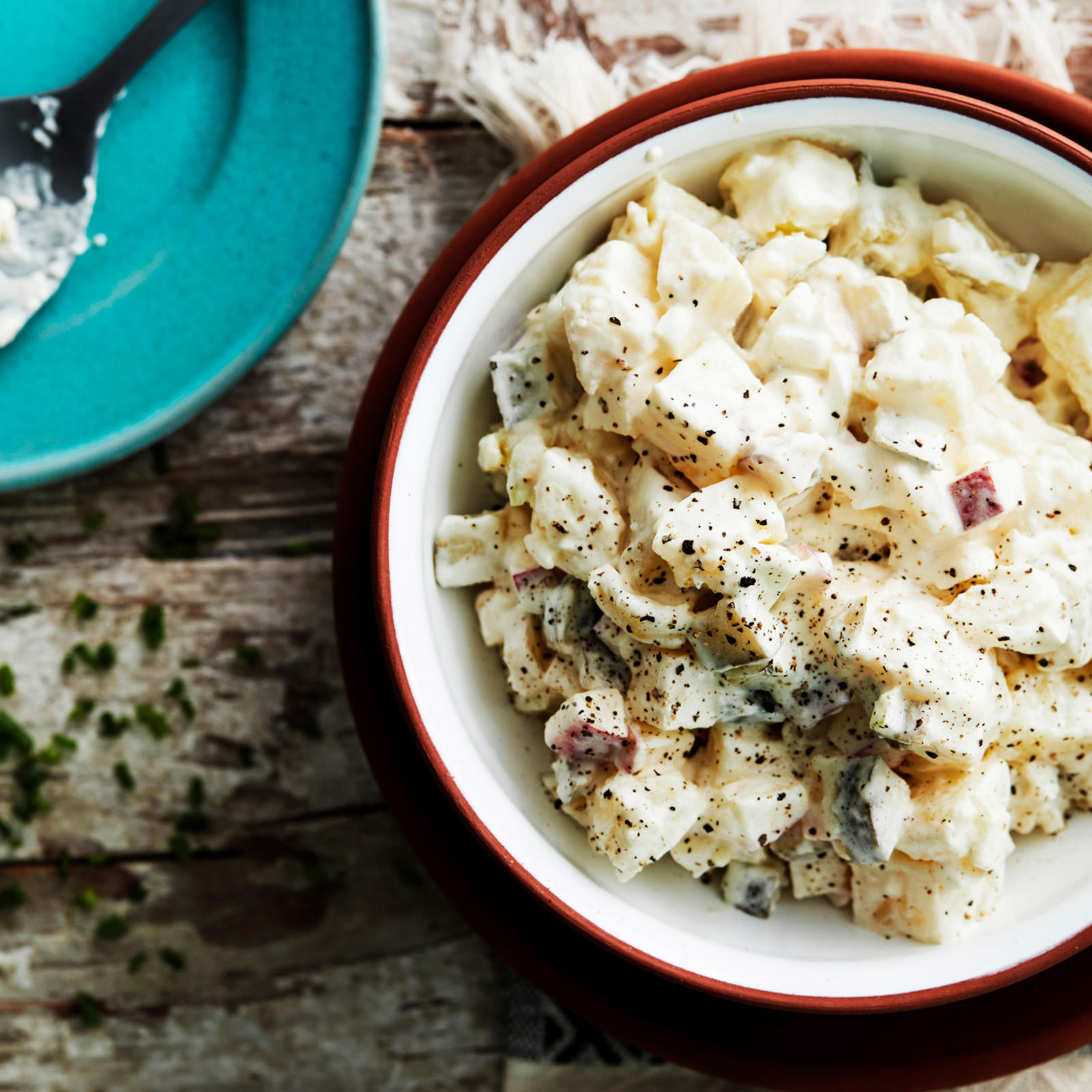 Perinteistä perunasalaattia kulhossa, jonka alla mustavalkoinen liina. Vieressä turkoosi lautanen, jonka päällä lusikka.