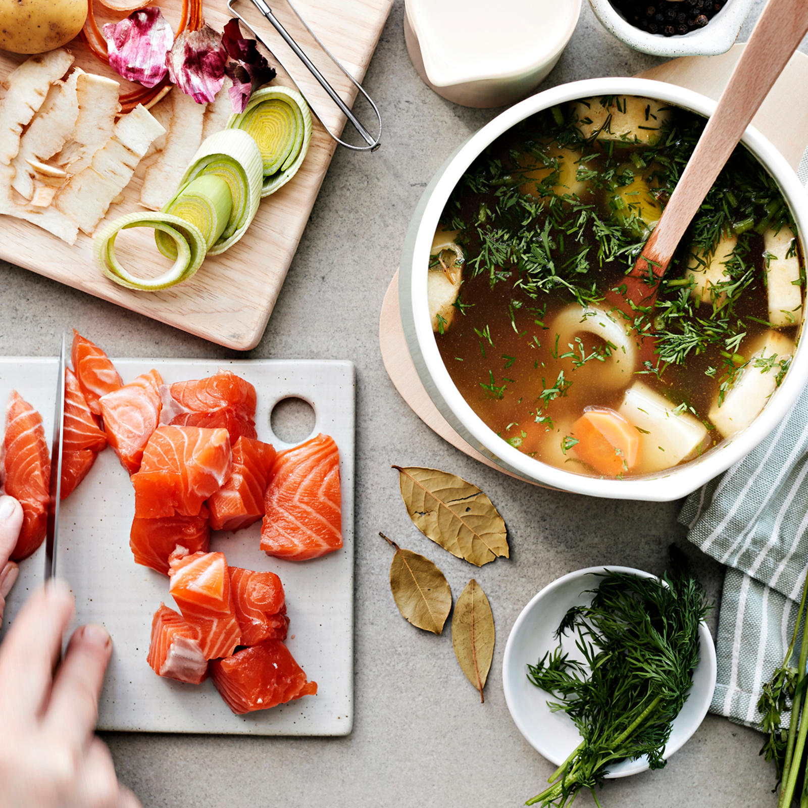 Kädet, jotka pilkkovat lohta paloiksi. Vieressä kasviksia ja keittokattila, jossa lientä ja kasviksia.