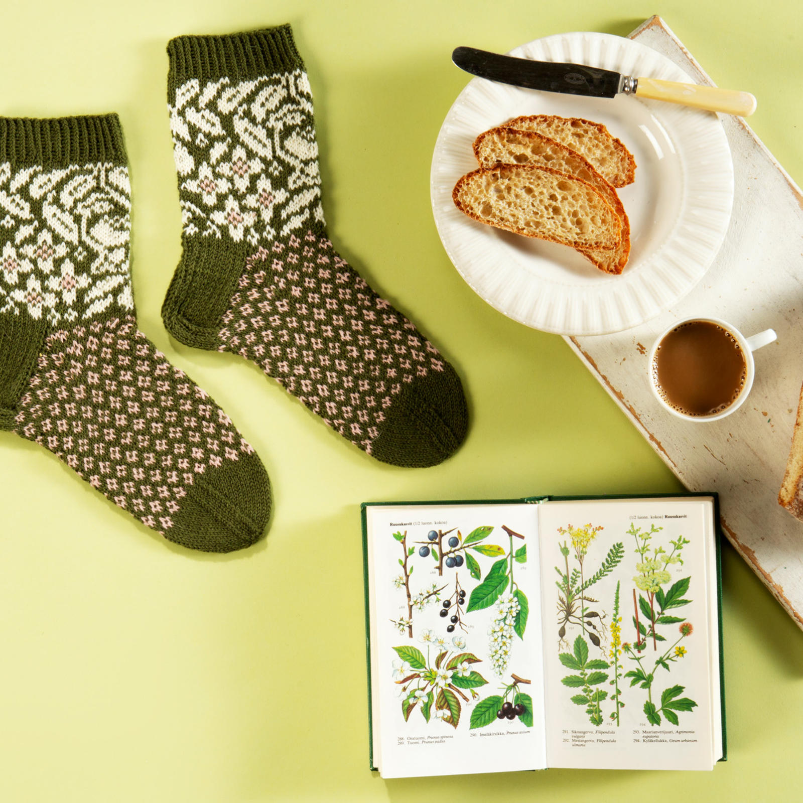 Villasukat, joissa on kukka- ja lintukuvioita. Somisteena kahvikuppi, kasvikirja ja leipä, josta on leikattu viipaleita lautaselle.