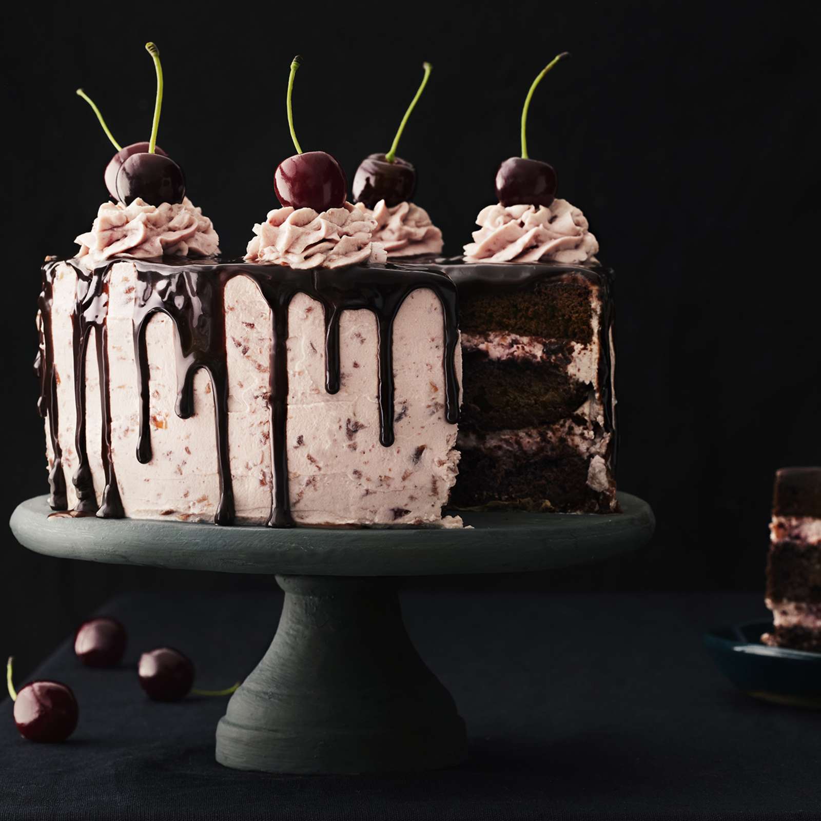 Tuoreilla kirsikoilla, pursotuksilla ja sulatetulla suklaalla koristeltu suklaa-kirsikkakakku tummalla, jalallisella kakkuvadilla. Kakusta leikattu pala lautaselle.
