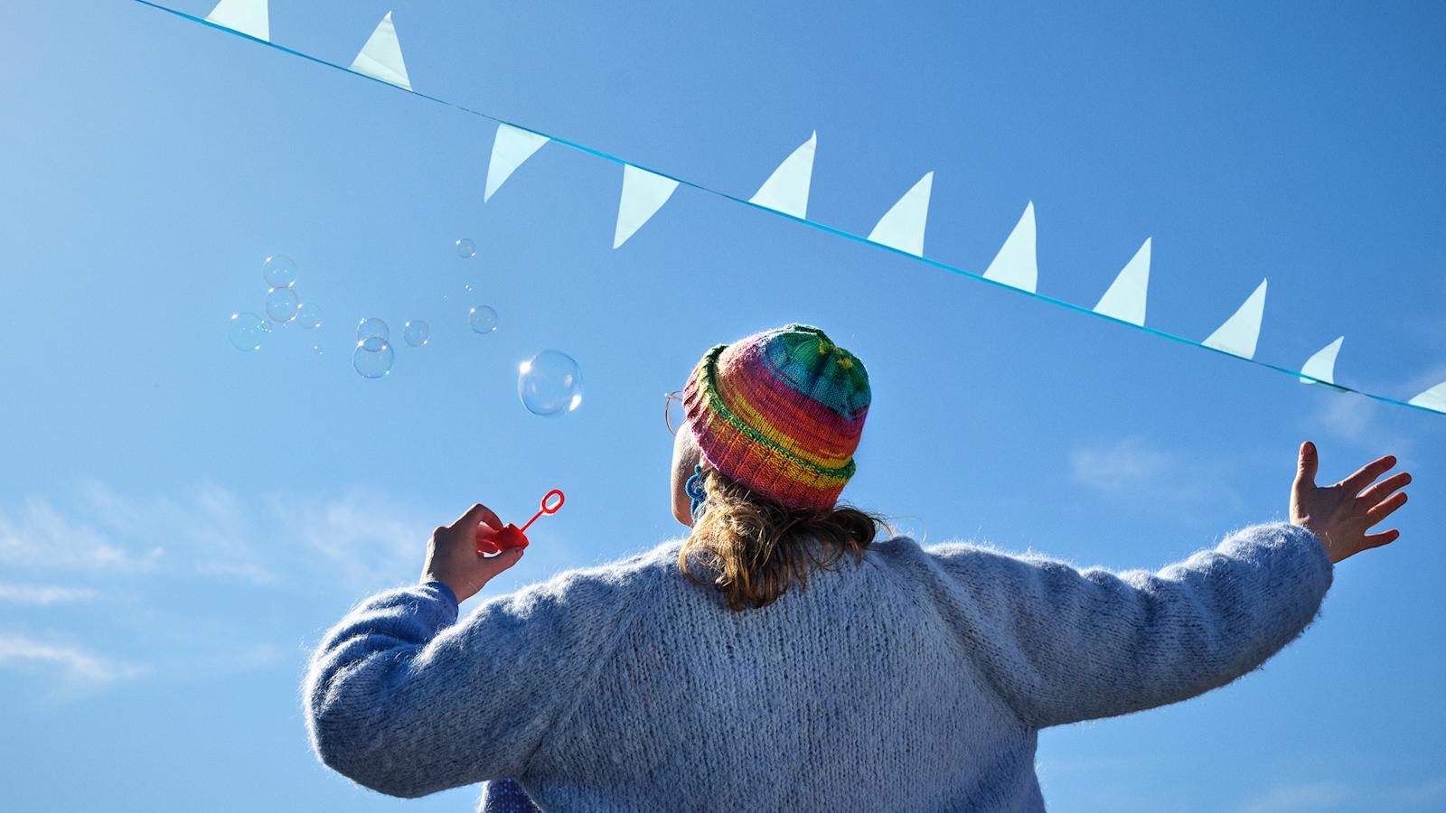 Sateenkaarenvärinen pipo päässään oleva henkilö, joka puhaltaa saippuakuplia.