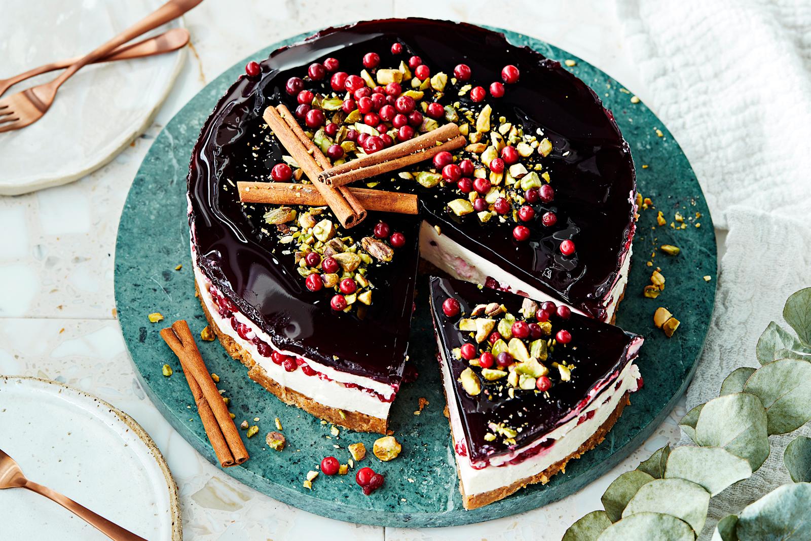 Glögijuustokakku, joka koristeltu kanelitangoilla, puolukoilla ja pistaaseilla. Kakusta leikattu yksi pala tarjoilulautaselle. Kakun vieressä lautasia ja leivoshaarukoita.