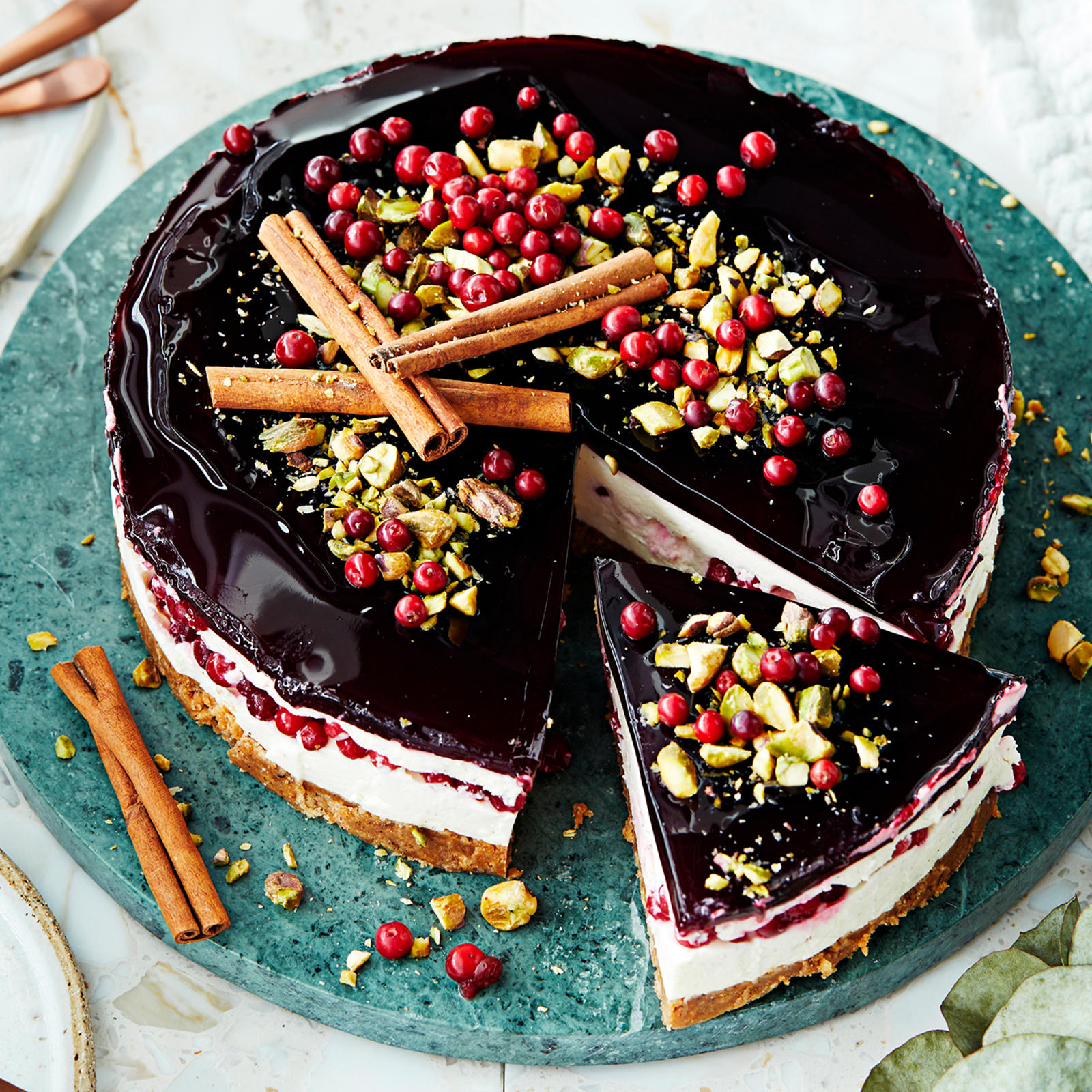 Glögijuustokakku, joka koristeltu kanelitangoilla, puolukoilla ja pistaaseilla. Kakusta leikattu yksi pala tarjoilulautaselle. Kakun vieressä lautasia ja leivoshaarukoita.