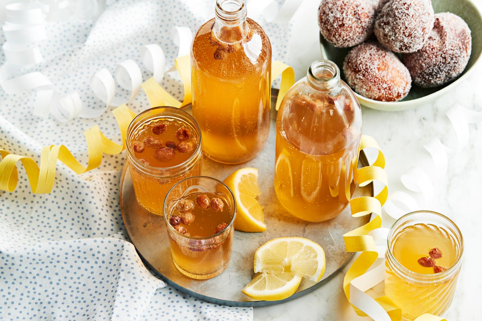 Simaa laseissa ja lasipulloissa tarjottimen päällä. Pilkullinen liina, serpentiiniä, sitruunaviipaleita sekä munkkeja kulhossa.