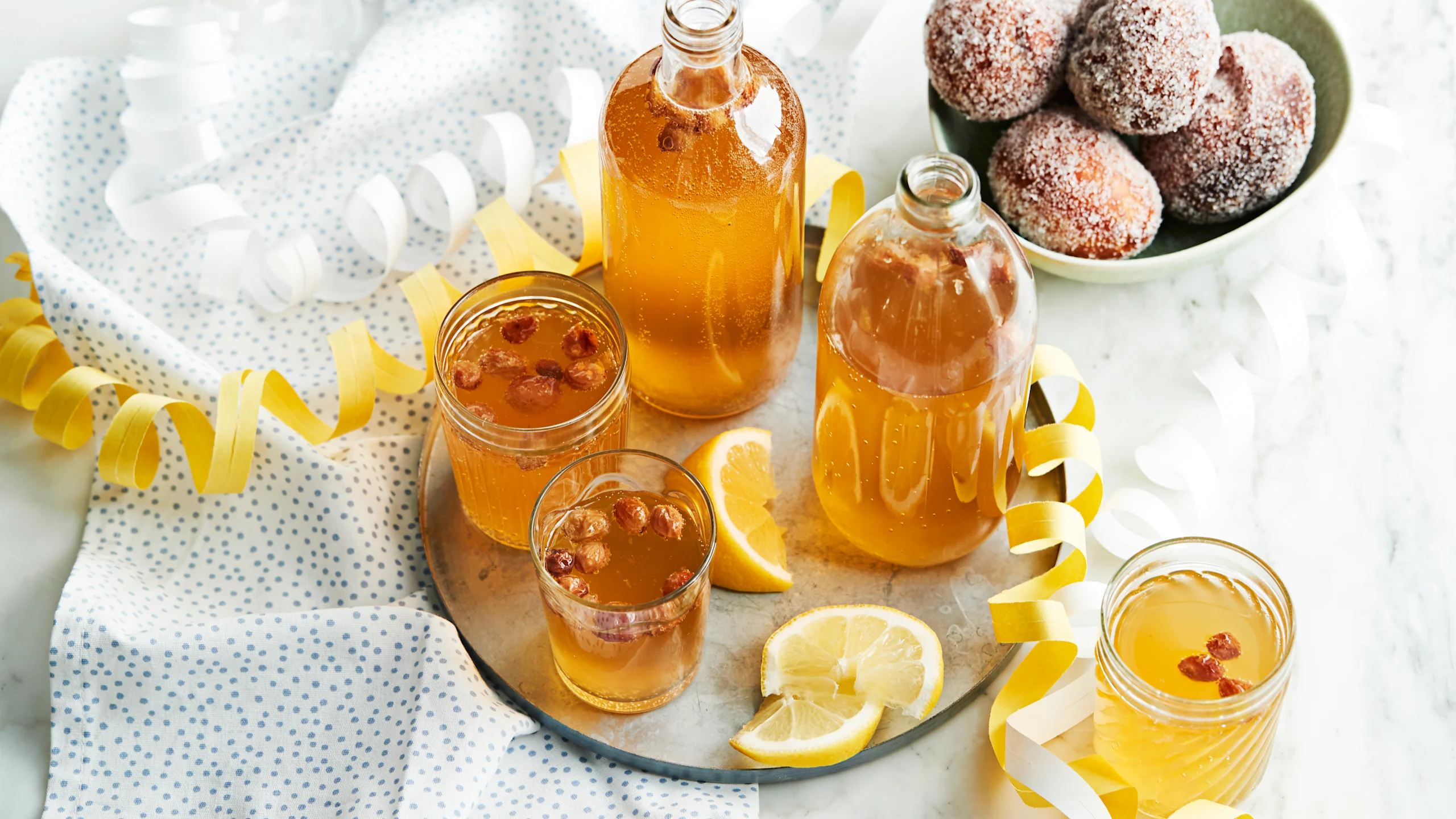 Simaa laseissa ja lasipulloissa tarjottimen päällä. Pilkullinen liina, serpentiiniä, sitruunaviipaleita sekä munkkeja kulhossa.
