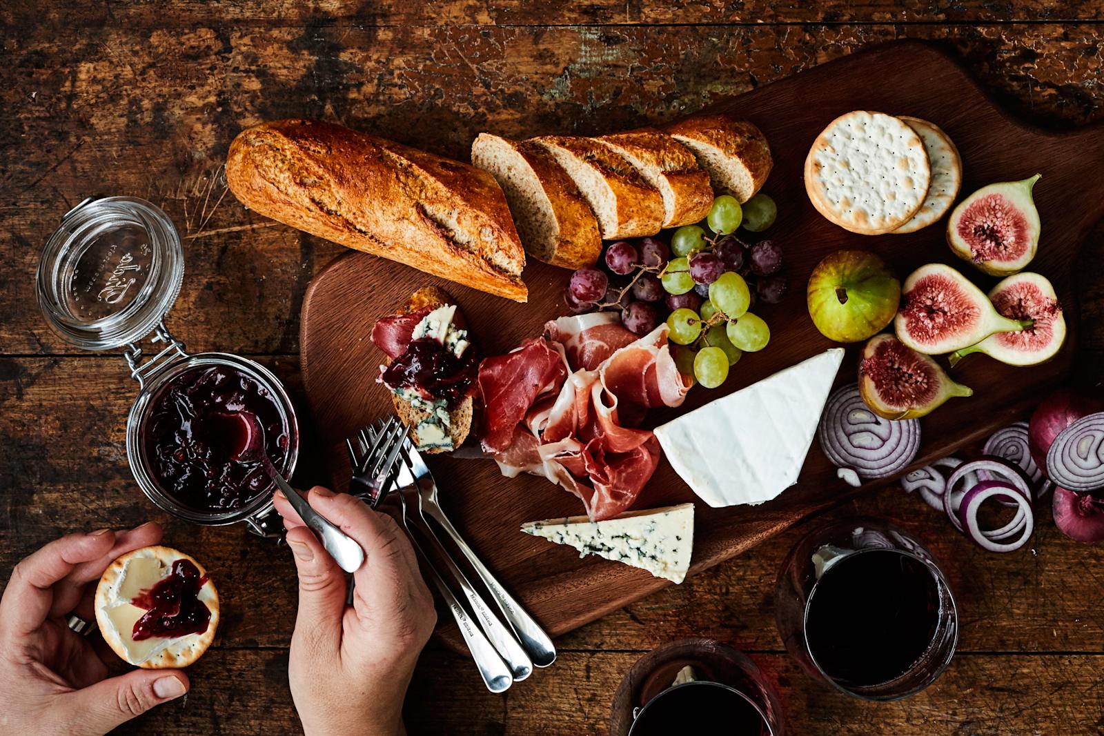 Punasipuli-puolukkahilloketta lasipurkissa puisella pöydällä. Vieressä tarjottimella juustoja, patonkia, leikkeleitä ja tuoreita hedelmiä.