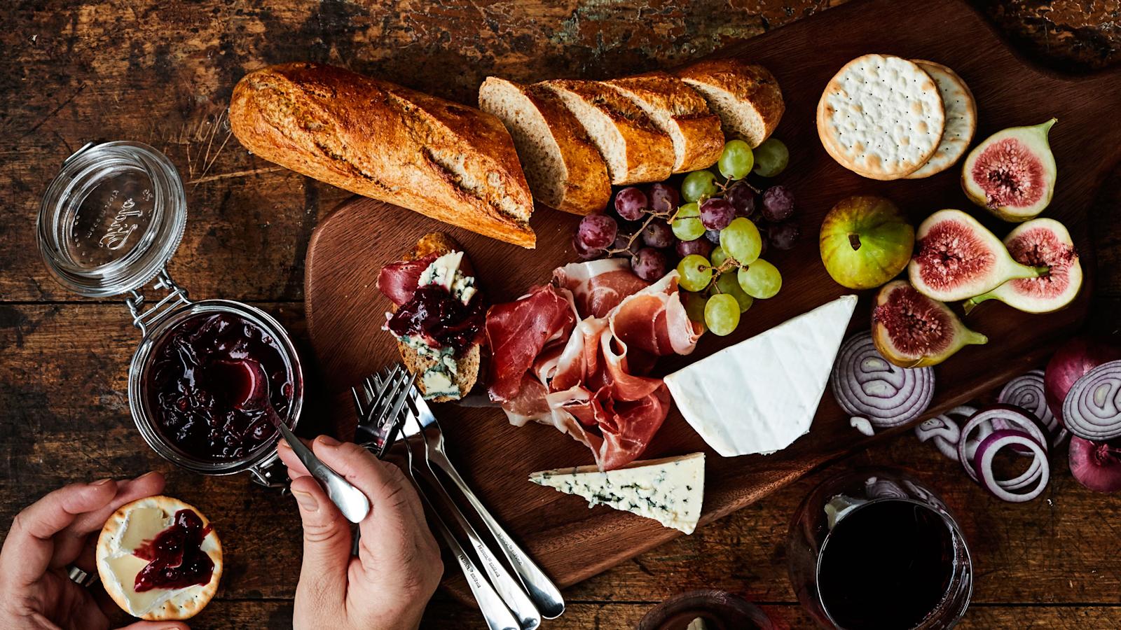 Punasipuli-puolukkahilloketta lasipurkissa puisella pöydällä. Vieressä tarjottimella juustoja, patonkia, leikkeleitä ja tuoreita hedelmiä.