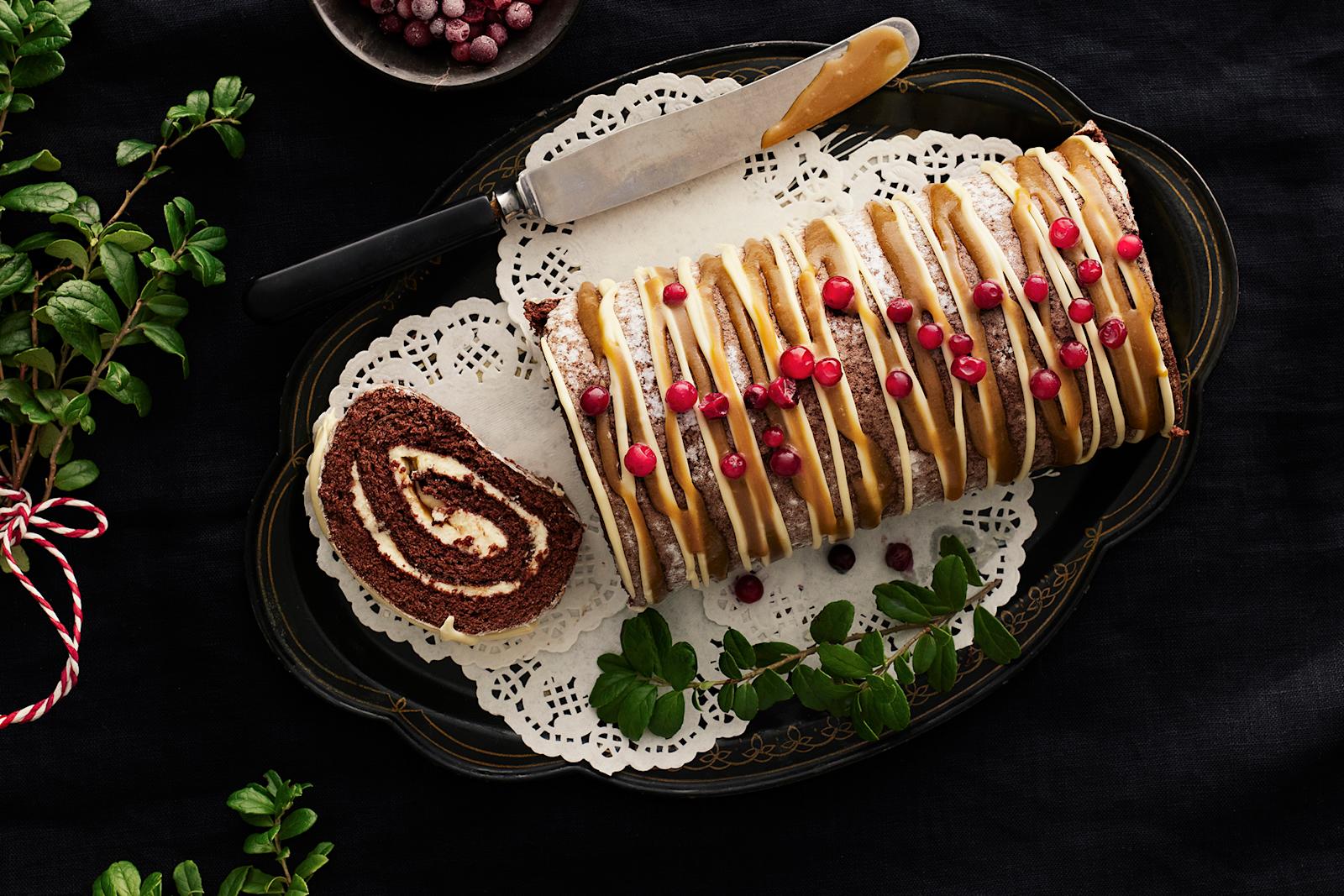 Valkosuklaa-kinuskijouluhalko, joka on koristelut puolukoilla. Leivonnaisesta leikattu viipale tarjoiluastialle. Tarjoiluastian reunalla veitsi ja vieressä puolukan varpuja.