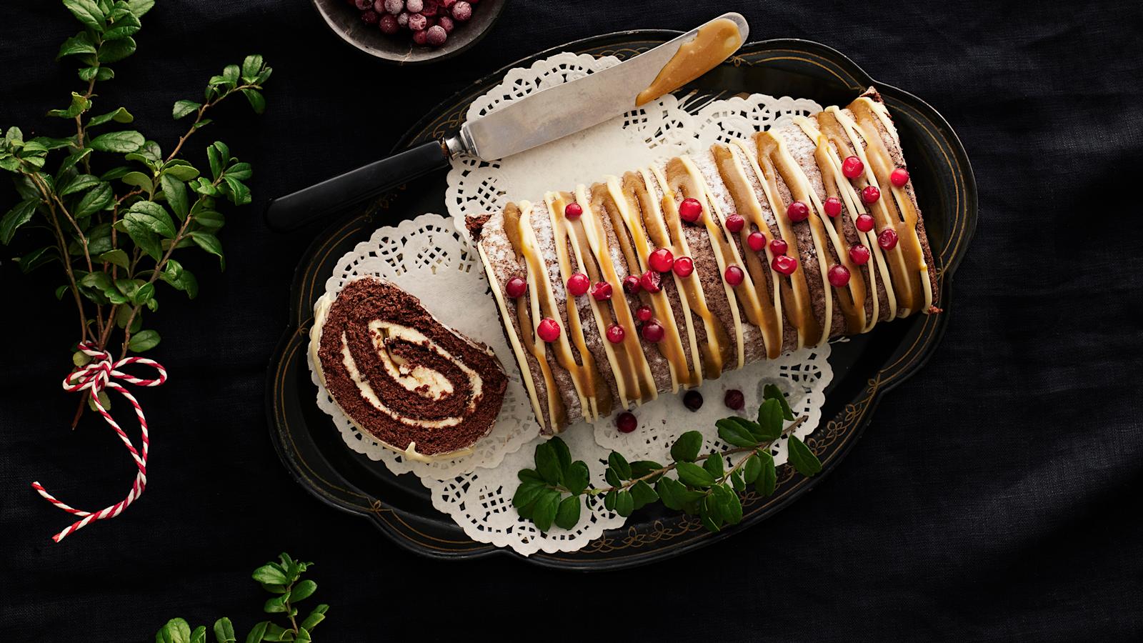 Valkosuklaa-kinuskijouluhalko, joka on koristelut puolukoilla. Leivonnaisesta leikattu viipale tarjoiluastialle. Tarjoiluastian reunalla veitsi ja vieressä puolukan varpuja.