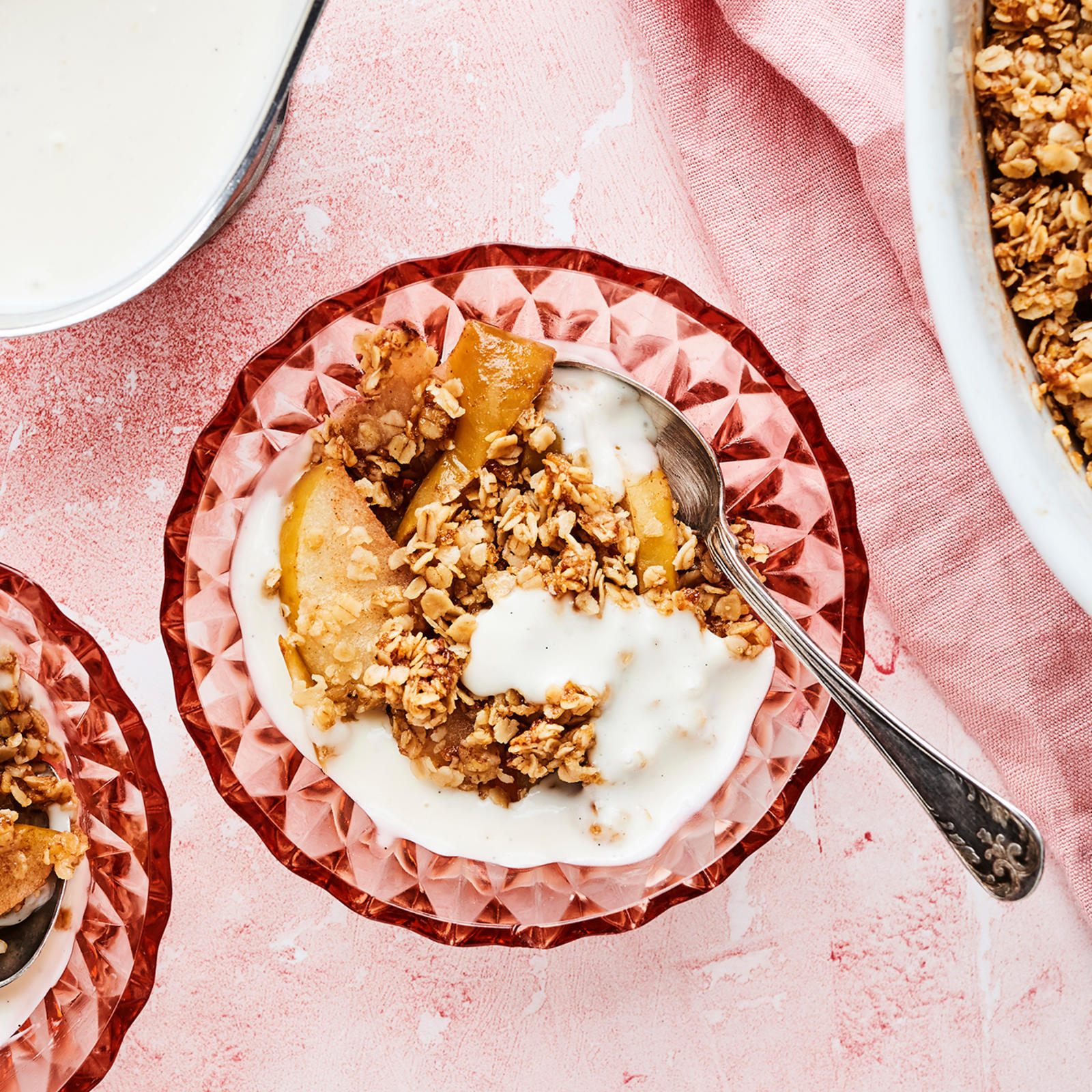 Kaura-omenapaistosta kahdessa lasikulhossa ja uunivuoassa. Uunivuoan alla vaaleanpunainen liina. Vanijakastiketta kastikekannussa.