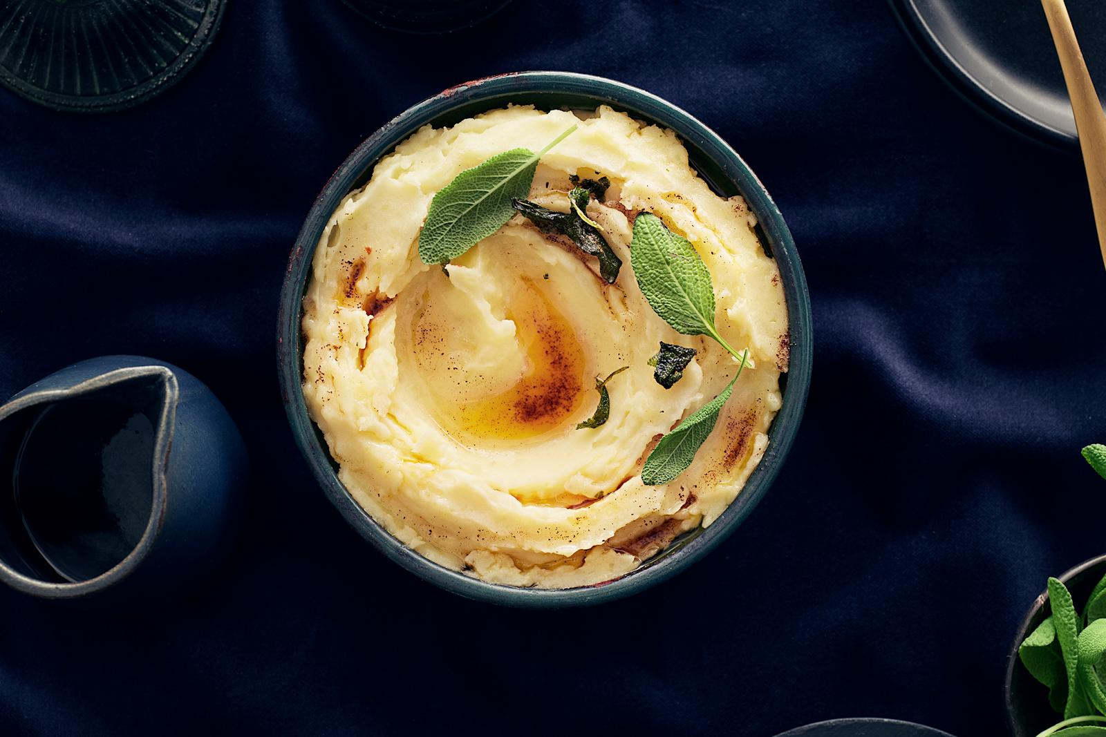 Kulhossa perunamuusia, jonka päällä salviavoita ja salvianlehtiä. Vieressä kastikekannu, jossa salviavoita.