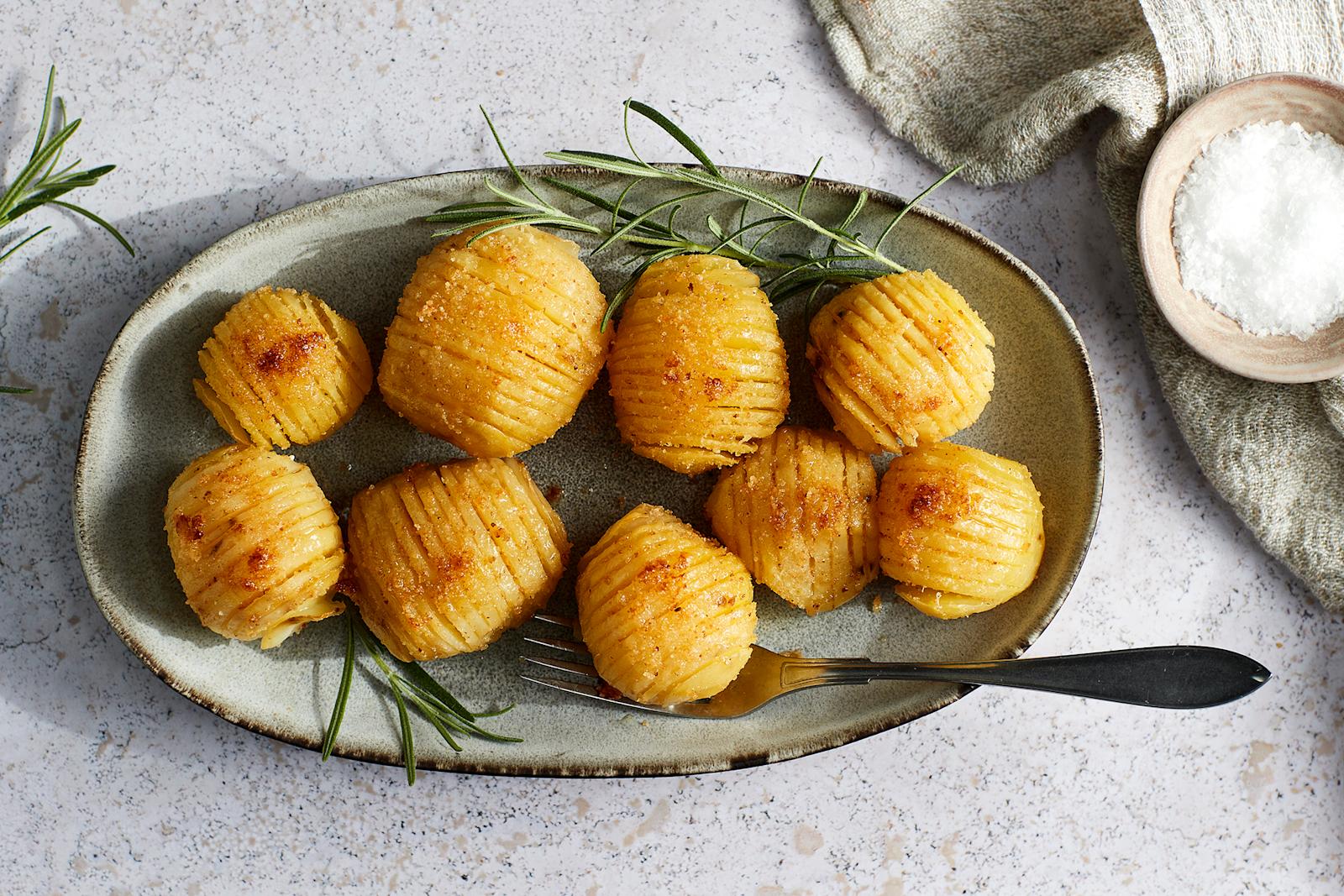 Hasselbackan perunat lautasella, jossa haarukka. Vieressä rosmariinin oksia, liina ja pieni kipollinen sormisuolaa.
