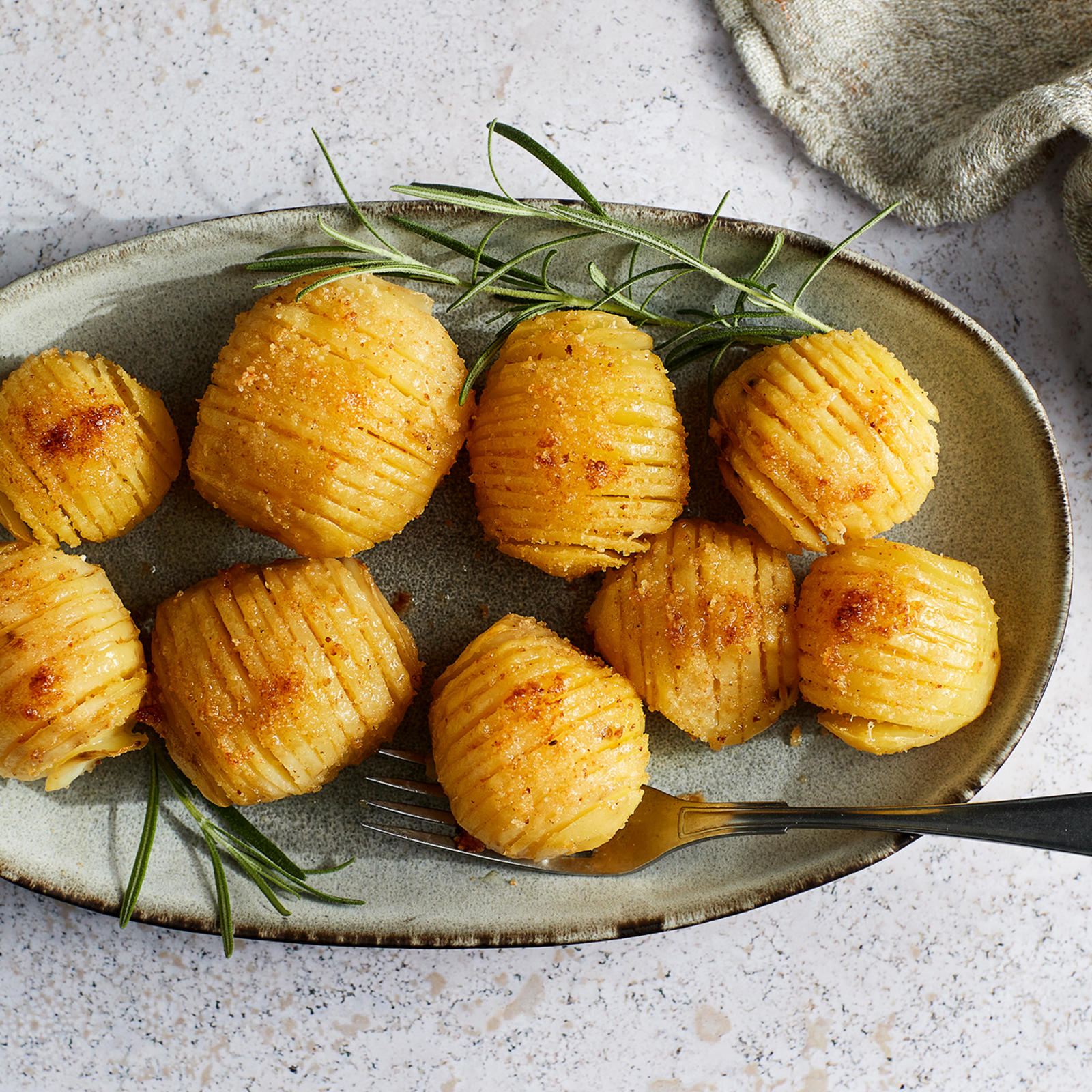 Hasselbackan perunat lautasella, jossa haarukka. Vieressä rosmariinin oksia, liina ja pieni kipollinen sormisuolaa.