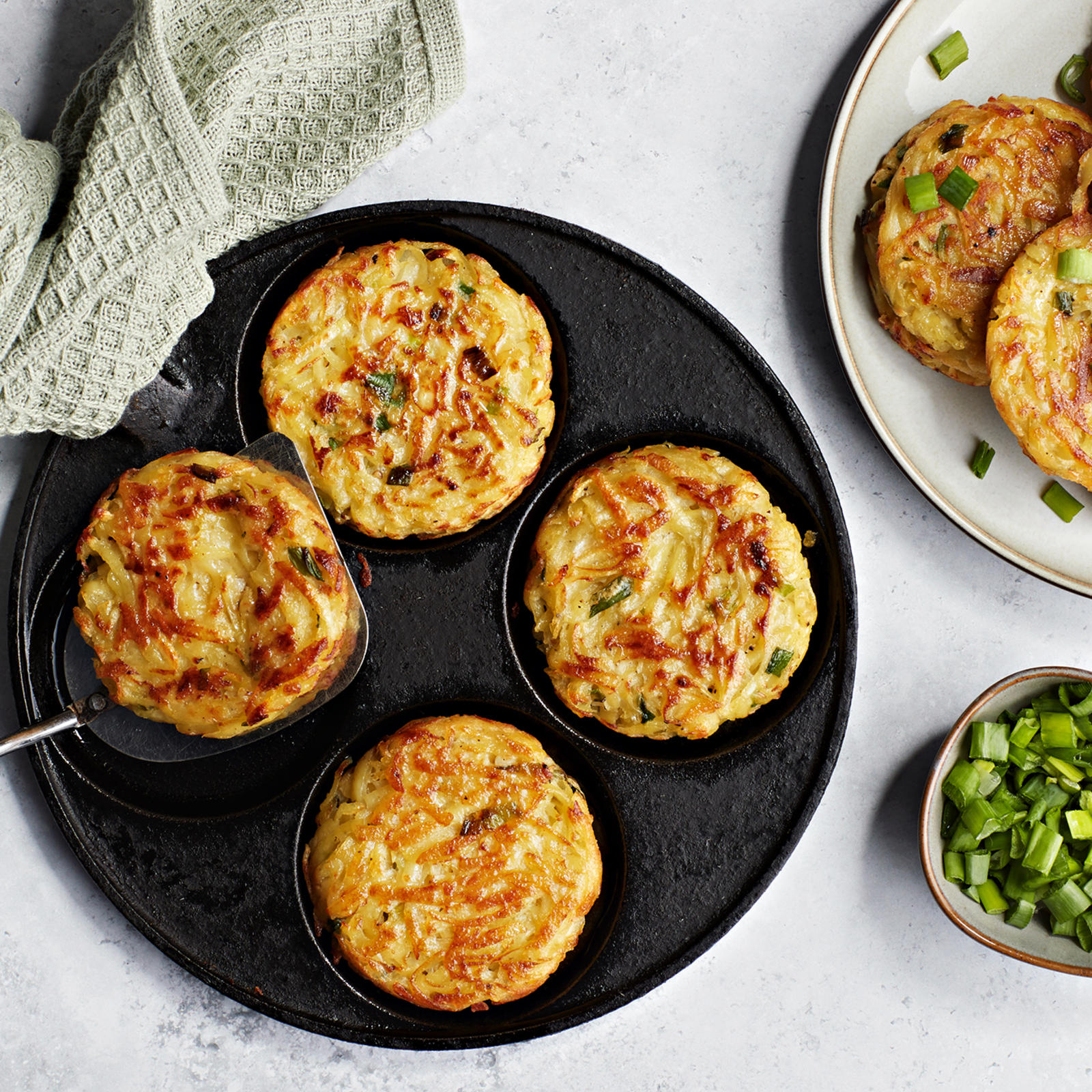 Röstiperunat ohukaispannulla. Vieressä valmiita röstiperunoita lautasella ja silputtua kevätsipulia kulhossa.
