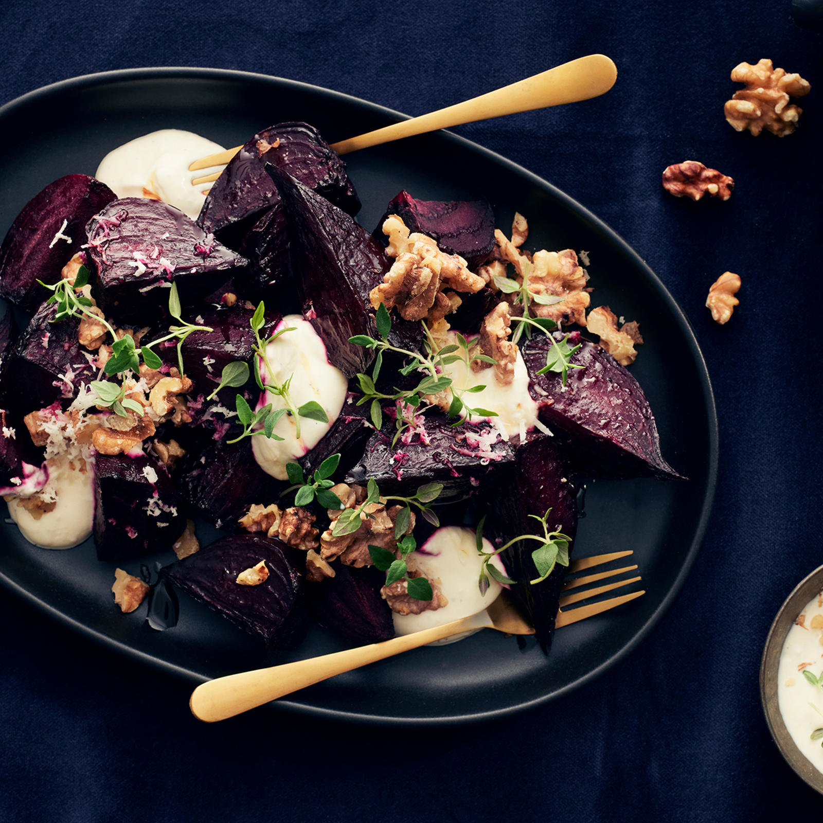 Paahdettuja punajuuria tummalla tarjoiluastialla piparjuurikastikkeella, saksanpähkinöillä ja timjamilla koristeltuna. Lautasella kolme haarukkaa. Lautasen vieressä saksanpähkinöitä ja piparjuurikastiketta kulhossa.