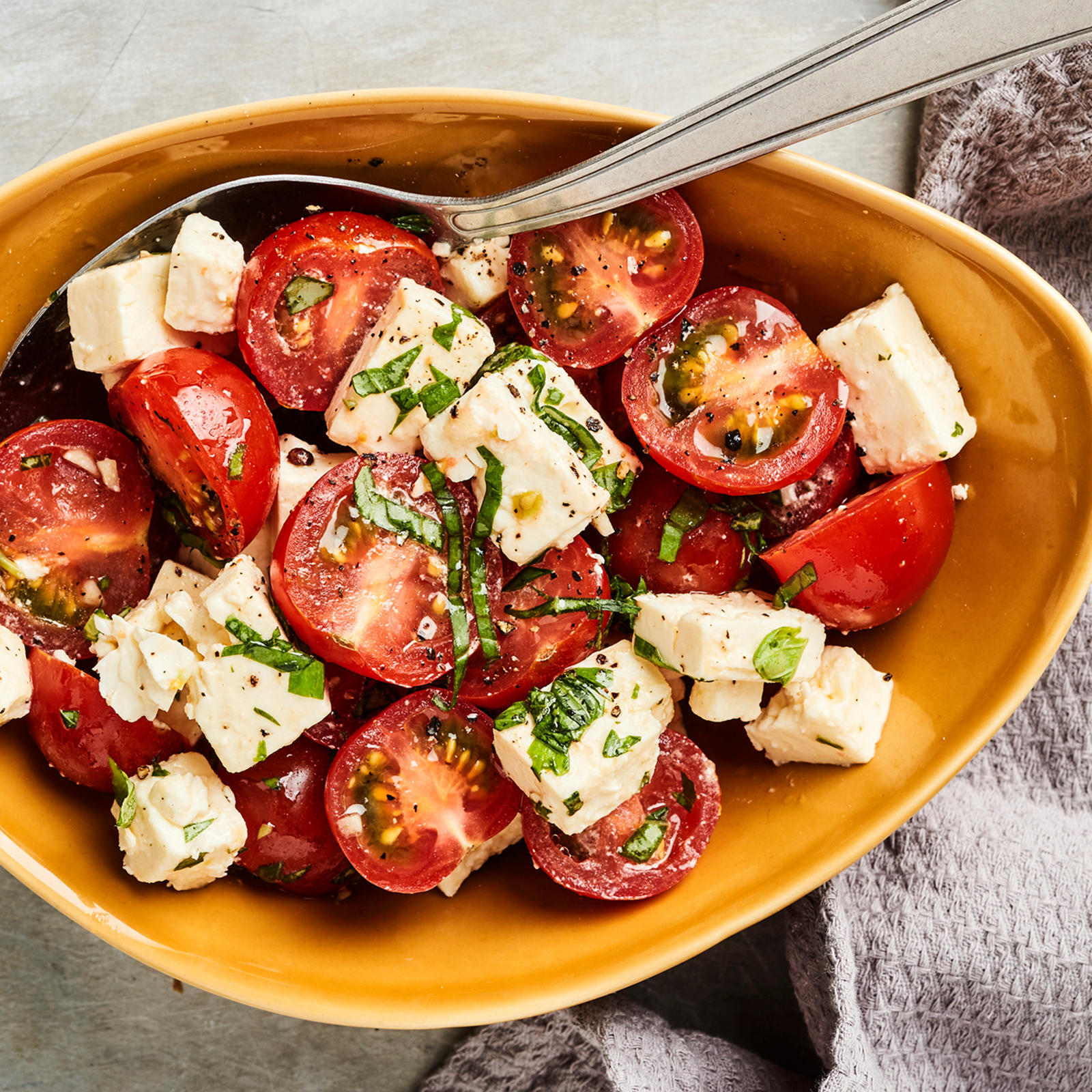 Marinoitu tomaatti-fetasalaatti keltaisessa kulhossa. Kulhossa lusikka ja kulhon vieressä harmaa liina.