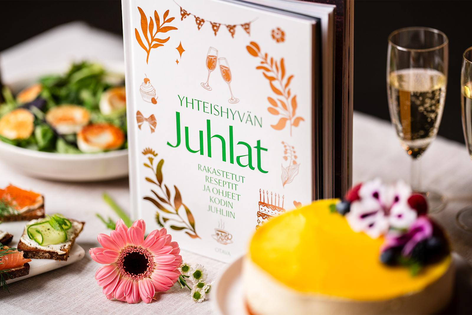 Yhteishyvän juhlat -kirja. Kirjan ympärillä juustokakku, voileipiä, salaattia ja kuohuviinilaseja.