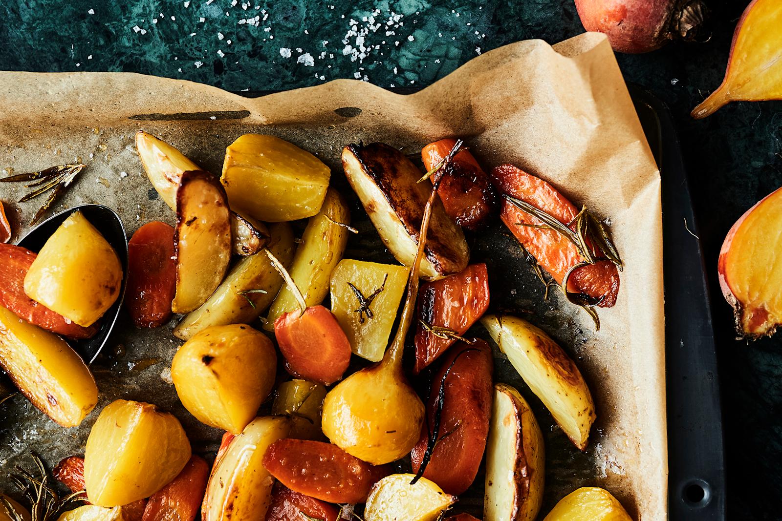 Leivinpaperilla päällystetyn uunipellin kulma, jolla uunijuureksia. Vieressä puolitettuja keltajuuria.