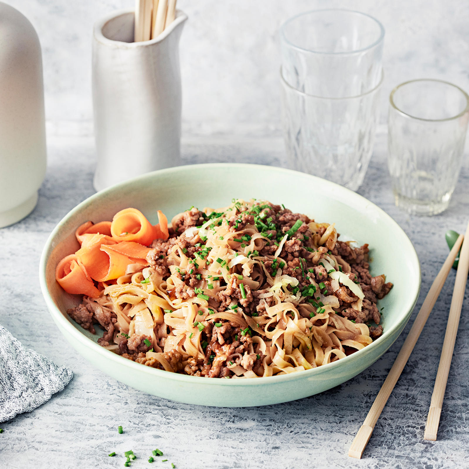 Dandan-nuudelit eli kiinalainen jauheliha-nuudelipannu lautasella. Vieressä syömäpuikot, laseja, liina ja kulhoissa porkkanasuikaleita ja hienonnettua ruohosipulia.