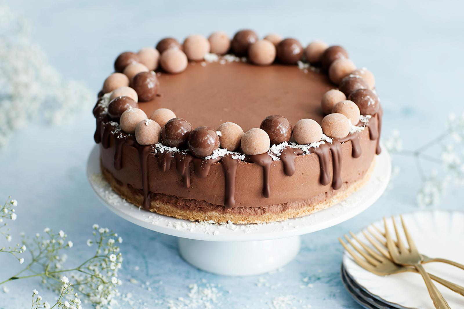 Suklaajuustokakku valkoisella kakkulautasella. Vieressä leivoshaarukoita, kakkulautasia ja harsokukkia.