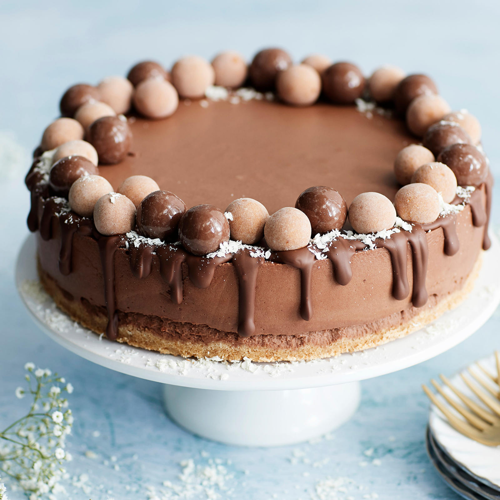 Suklaajuustokakku valkoisella kakkulautasella. Vieressä leivoshaarukoita, kakkulautasia ja harsokukkia.