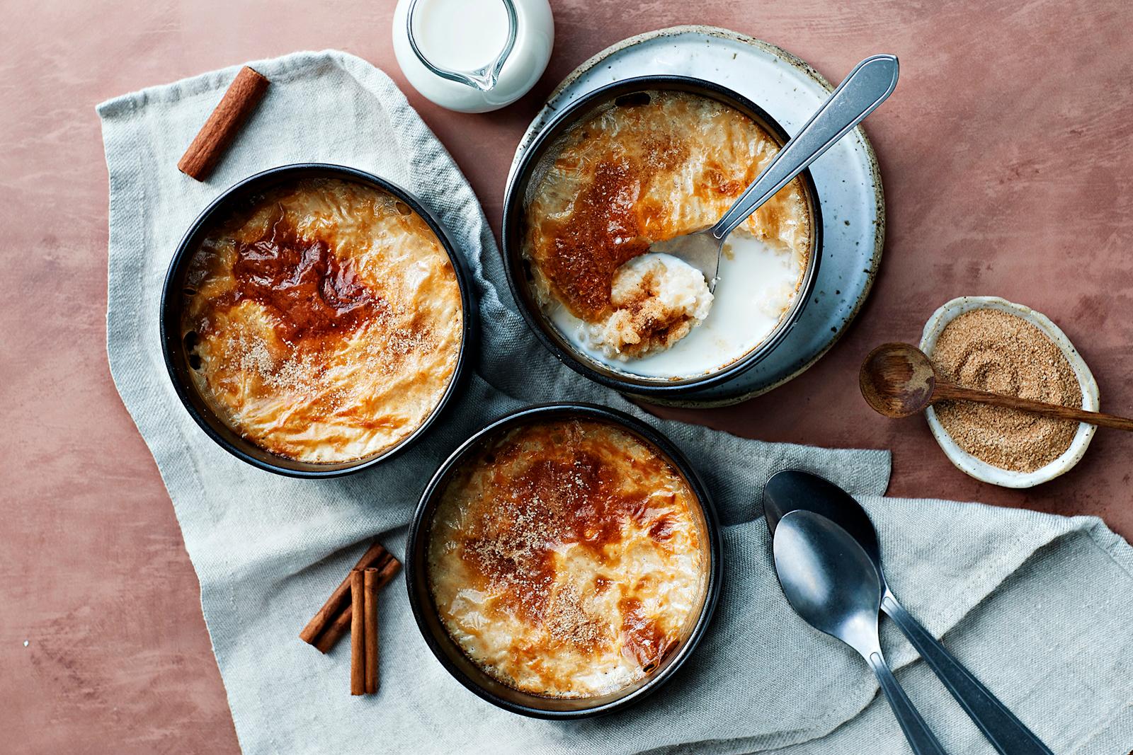 Uuniriisipuuro valmistettuna kolmessa kulhossa. Kulhojen alla liina ja vieressä lusikoita, kanelisokeria ja maitokannu.