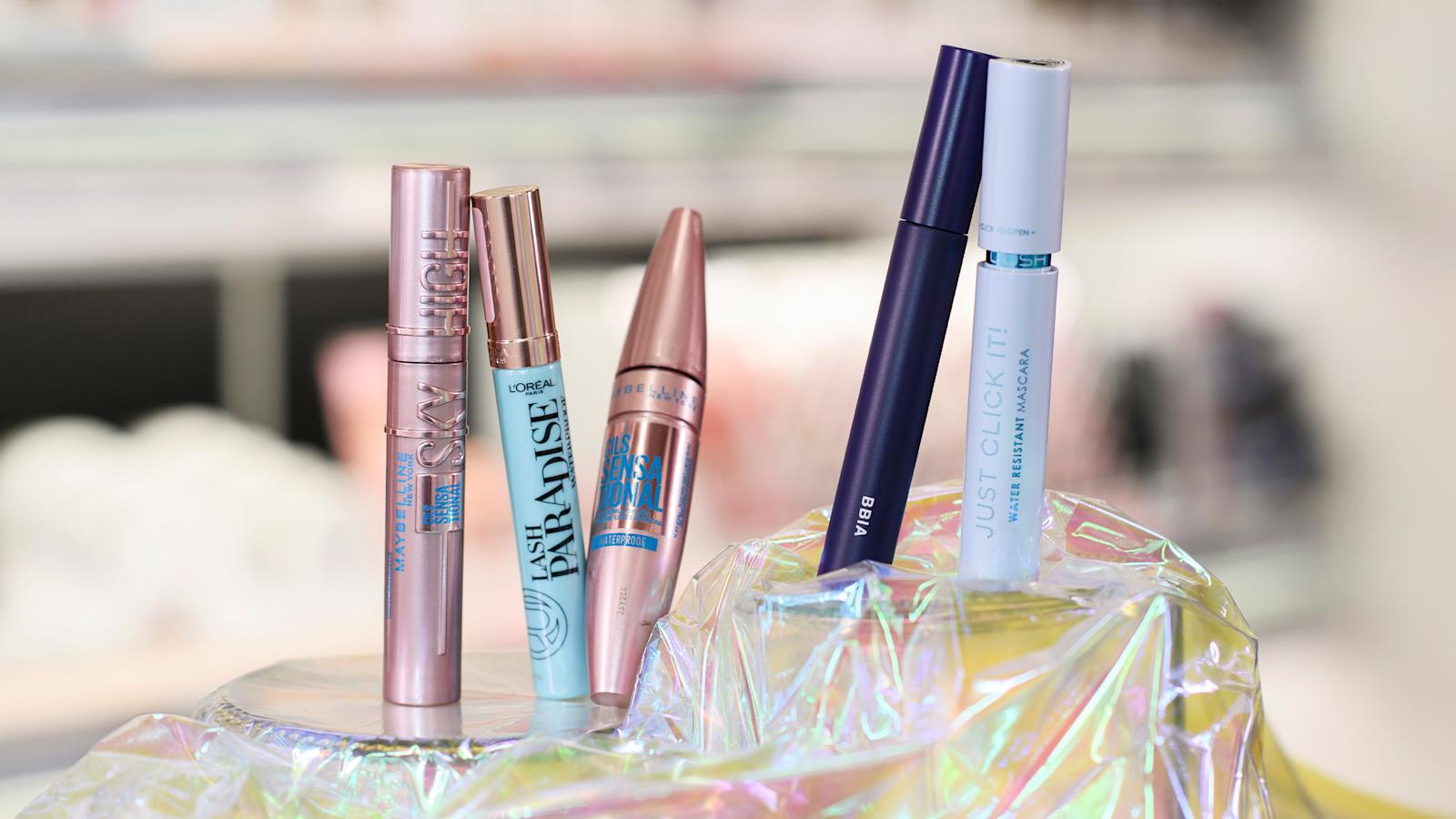 Viisi erilaista ripsiväriä kuvattuna hohtavan kankaan päällä, taustalla kaupan meikkihylly.