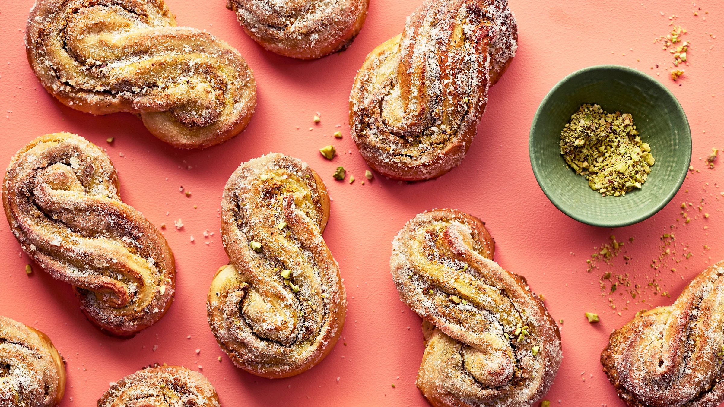 Ässäpullia oranssilla taustalla. Vieressä vihreä kulho, jossa rouhittuja pistaasipähkinöitä.