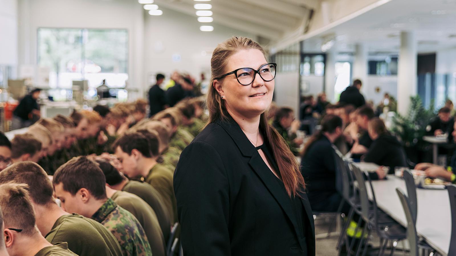 Nainen seisoo ruokalassa, jossa armeijaa käyvät nuoret ruokailevat