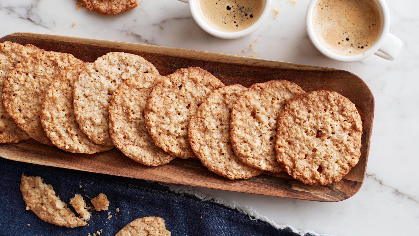 Kaurakeksejä puisella tarjoilulautasella. Lautasen vieressä kaksi kahvimukia ja tumma liina, jonka päällä murusteltuja keksejä.