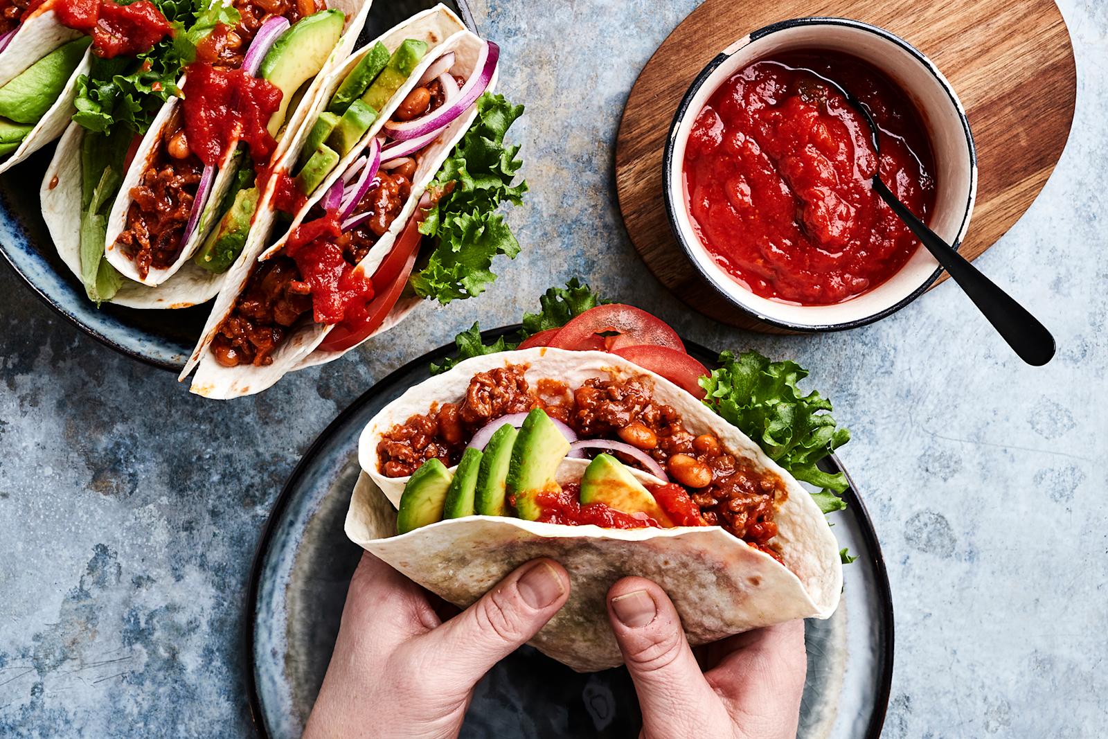 Kädet, jotka pitelevät jauheliha-papuseoksella ja kasviksilla täytettyä tortillaa. Vieressä kulho, jossa salsaa sekä valmiiksi täytettyjä tortilloja.