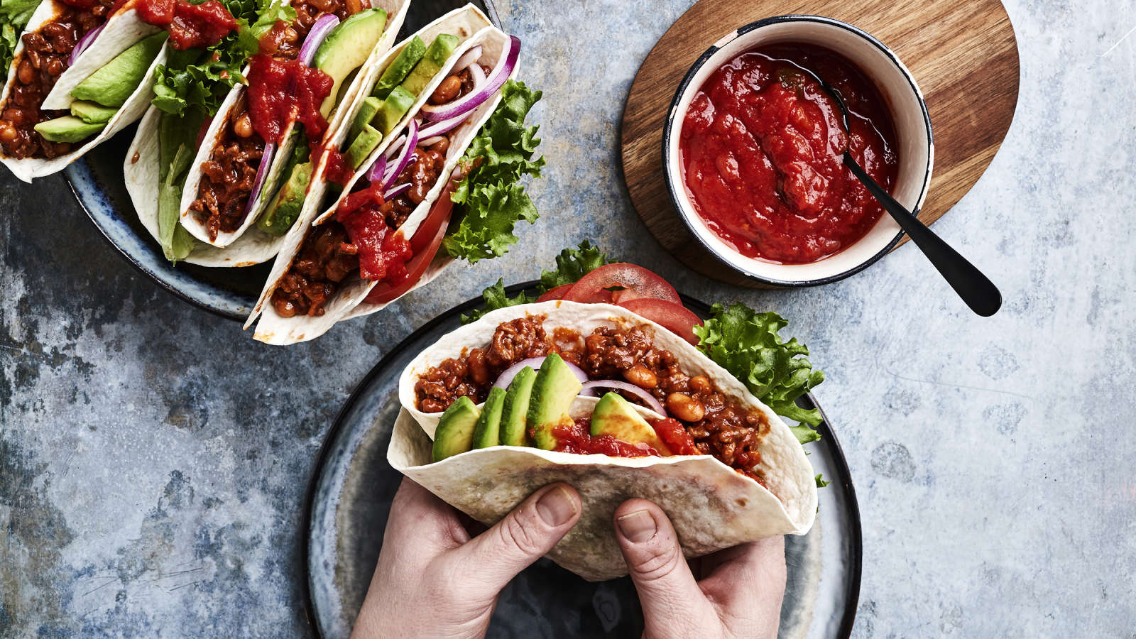 Kädet, jotka pitelevät jauheliha-papuseoksella ja kasviksilla täytettyä tortillaa. Vieressä kulho, jossa salsaa sekä valmiiksi täytettyjä tortilloja.